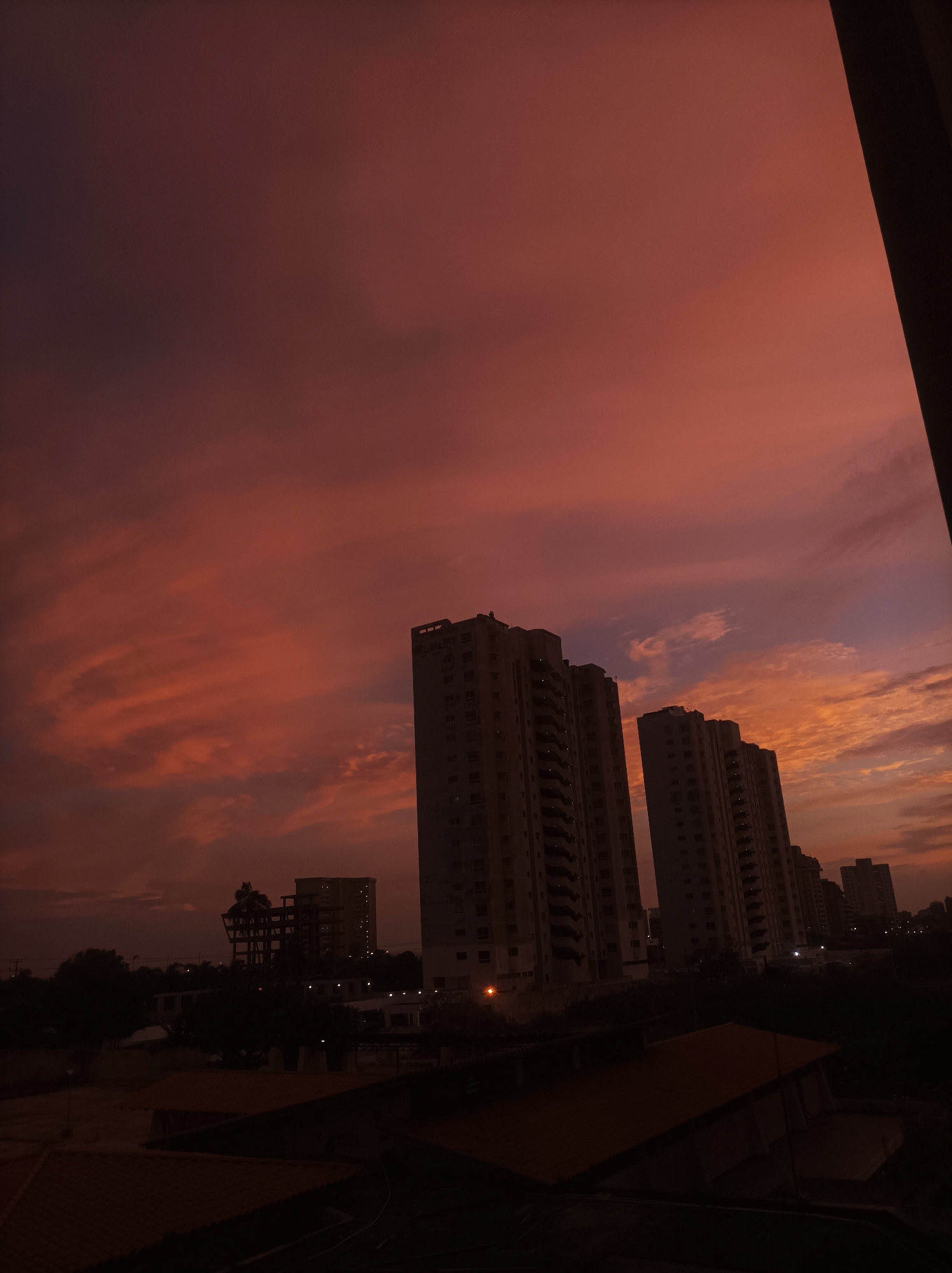 The image depicts a cityscape at dusk in Porlamar, Venezuela. Several tall, light-colored apartment buildings dominate the midground, silhouetted against a vibrant, reddish-orange sky with streaks of clouds. In the foreground are the dark rooftops of smaller buildings or structures, partially obscuring the view of the city below. The sky is the most prominent feature, displaying a blend of warm hues indicative of sunset. There are no people visible in the frame. The time of day is near sunset.