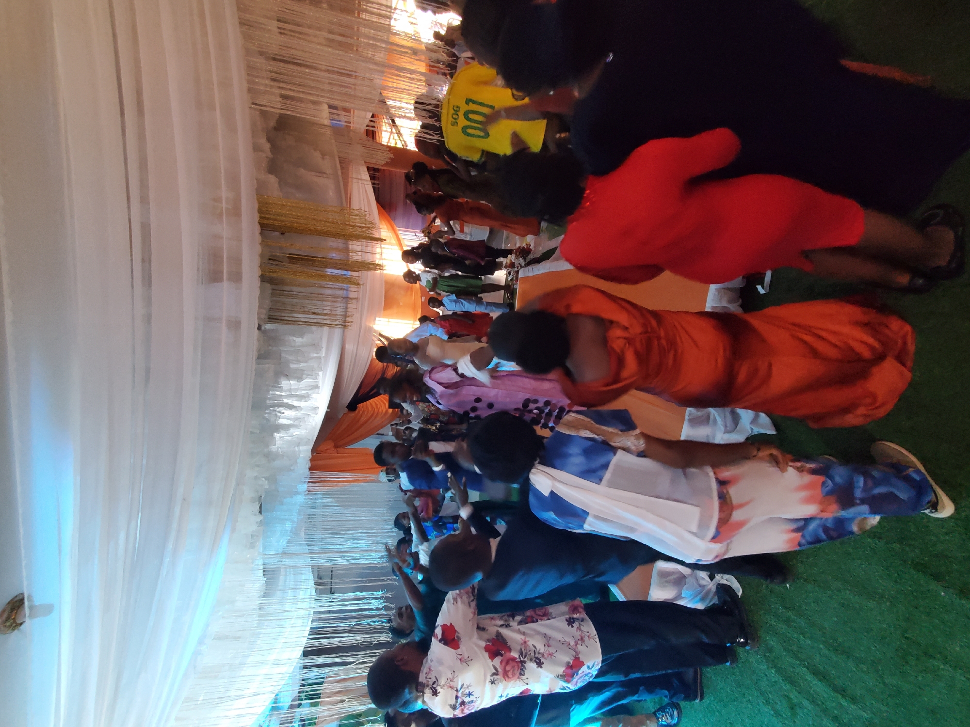 The image shows a gathering of people in an indoor setting, possibly a hall or event space. The scene appears to be a celebration or event of some kind, given the decorations like the white drapes and golden string decorations overhead. The people are dressed in various attire, including a yellow shirt with "007 SOG" printed on it, a red dress, and a multi-colored patterned top. Many are seated while others are standing. Some individuals are interacting or focused on a point of interest within the event space. The location is Unknown, Unknown.