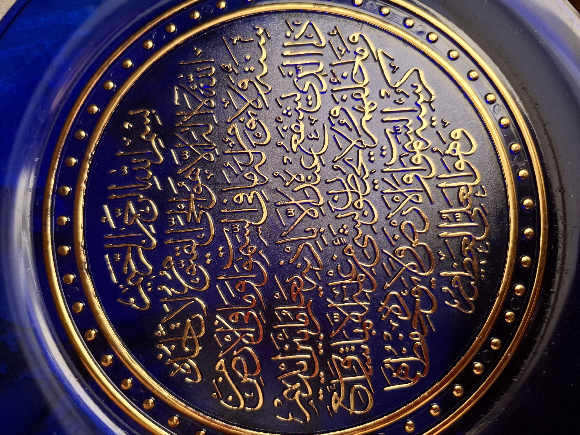 The image depicts a round, blue plate adorned with intricate gold-colored calligraphy. The calligraphy is densely packed within the central circular area of the plate. The plate is edged with a thin, gold-colored ring punctuated by evenly spaced, small, round, gold-colored embellishments. The lighting suggests a direct light source, creating highlights and shadows on the plate's surface and enhancing the depth of the calligraphy. The plate appears to be resting on a surface with an undefined background. The image does not provide any information about the city of Амзя, Russia, or its location.