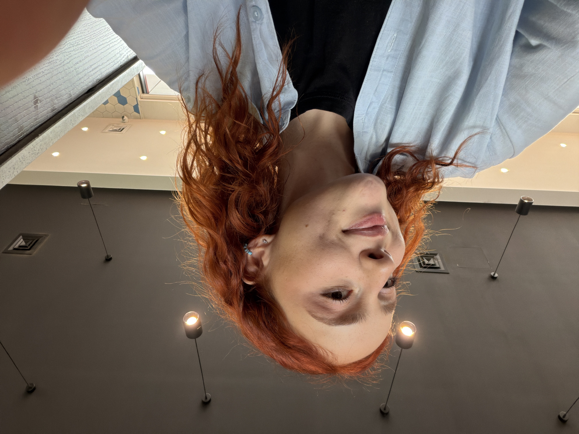 The media file features a close-up, low-angle shot of a young individual with reddish-brown, curly hair. The person is positioned upside down in the frame, looking up towards the viewer with a gentle, soft smile. They have fair skin, a nose piercing, and a small blue earring in their upper ear cartilage. They are wearing a light blue, collared button-up shirt over a black t-shirt.
The activity captured is a selfie taken from a worm's-eye view, making the subject appear as if they are looking down at the camera. The setting is indoors, with a dark grey ceiling visible in the background. Several bright, modern light fixtures are scattered across the ceiling: some are recessed spotlights, while others are hanging pendant lights with glowing bulbs. A rectangular air vent or grille is also visible on the ceiling.
Notable details include the subject's distinct hair color and curly texture, the subtle smile, and the unusual perspective of the shot. One cropped hint reveals a bright area above the person, showing blue and white hexagonal tiles on what appears to be an upper wall or ceiling, possibly indicating a restroom or a decorative architectural feature. No text is visible in the image. The time of day appears to be well-lit, likely indoors. While the location context of Tbilisi, Georgia, is provided, it is not visually evident in the image itself.