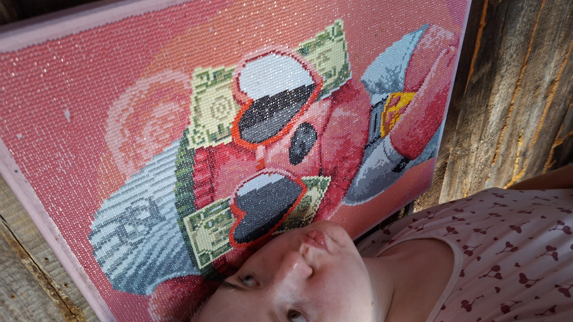 The image shows a woman holding a diamond painting. The diamond painting depicts a red car with sunglasses and money. The woman is lying on what appears to be a wooden deck. The location is Большой Букор, Russia.