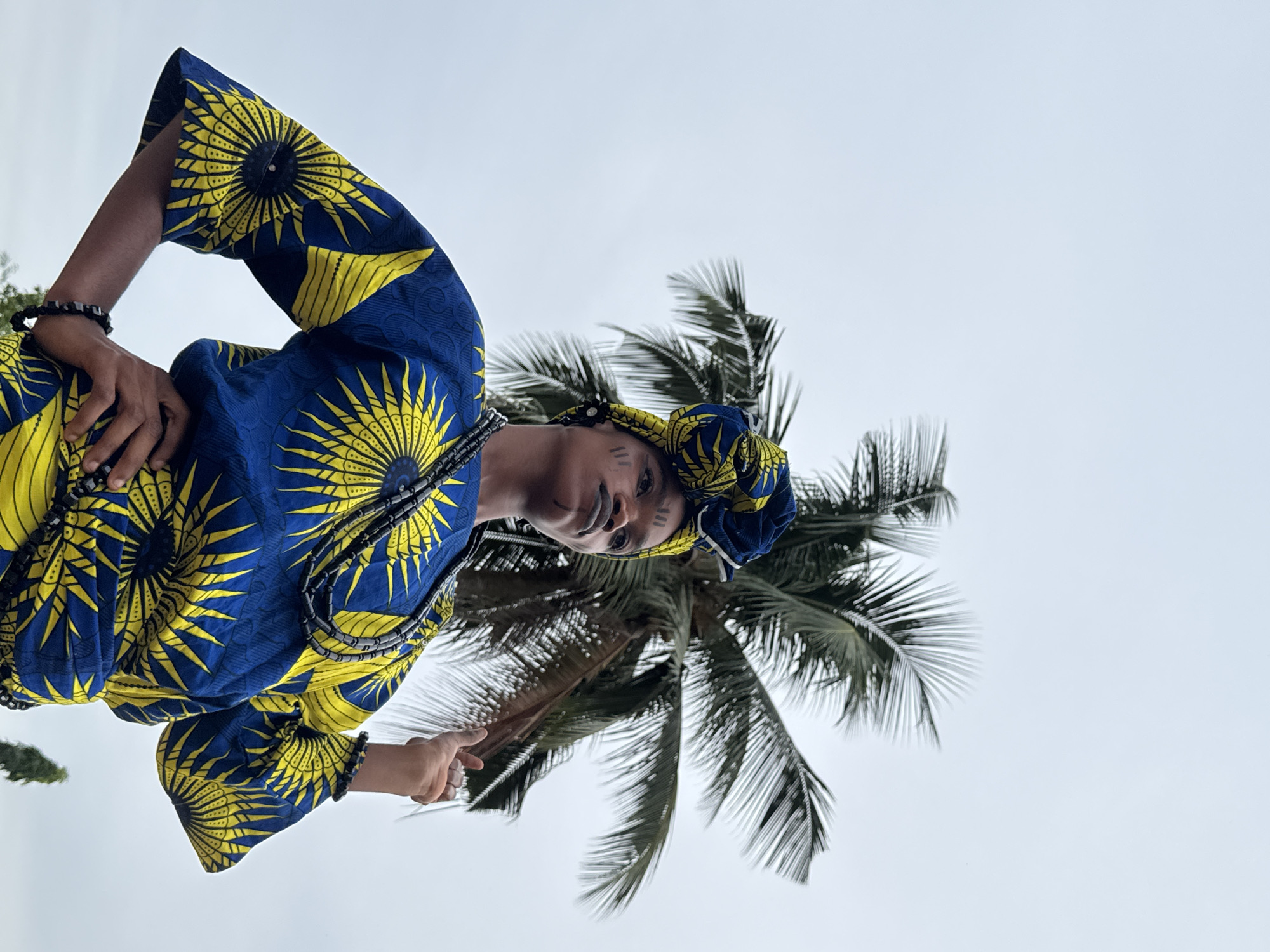 A woman stands next to a coconut palm in Badagry, Nigeria. She wears a blue and yellow printed dress and a matching headwrap, along with beaded necklaces and bracelets. Her face is adorned with white paint markings. The background consists of a pale sky and the fronds of the palm tree.