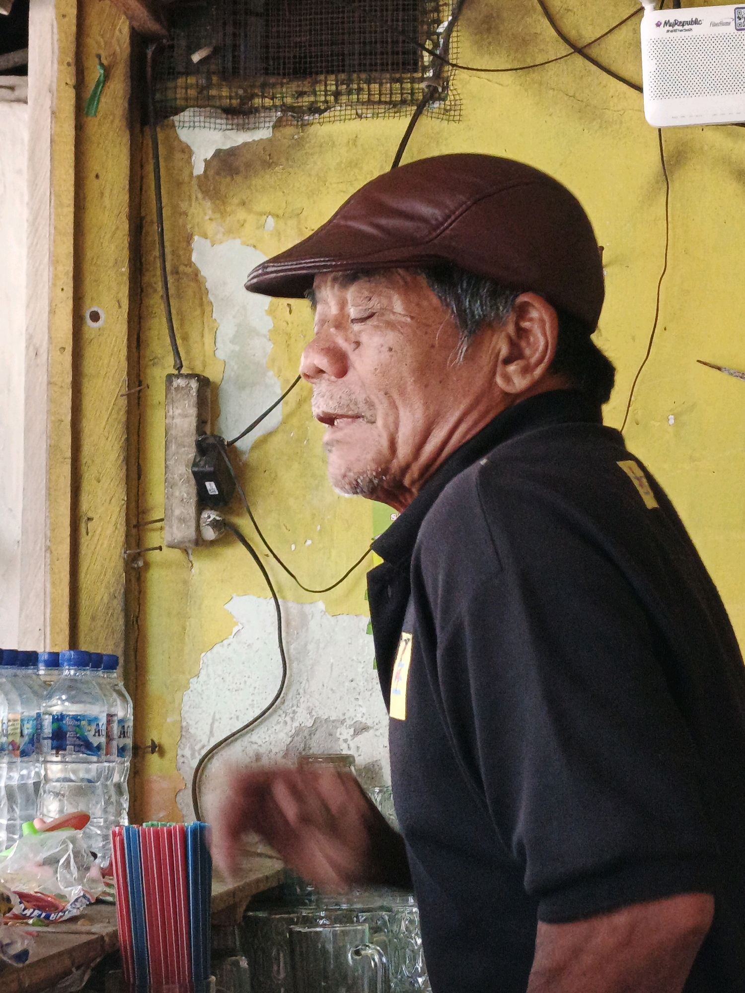 The image depicts a middle-aged man wearing a brown flat cap and a dark shirt, standing indoors. He is facing to the right, with his eyes closed and his mouth slightly open. His skin appears weathered. Behind him, a yellow wall exhibits peeling paint and exposed plaster, with electrical wires and a primitive-looking electrical outlet mounted on a concrete block. To the man's left, a stack of plastic water bottles is visible, along with a bundle of red and blue plastic straws. In the foreground, partially obscured by motion blur, is a hand reaching towards a surface where several glasses are present. The scene appears to be in a modest, possibly outdoor, establishment given the materials and state of the wall. The location is identified as Ketegan, Indonesia.