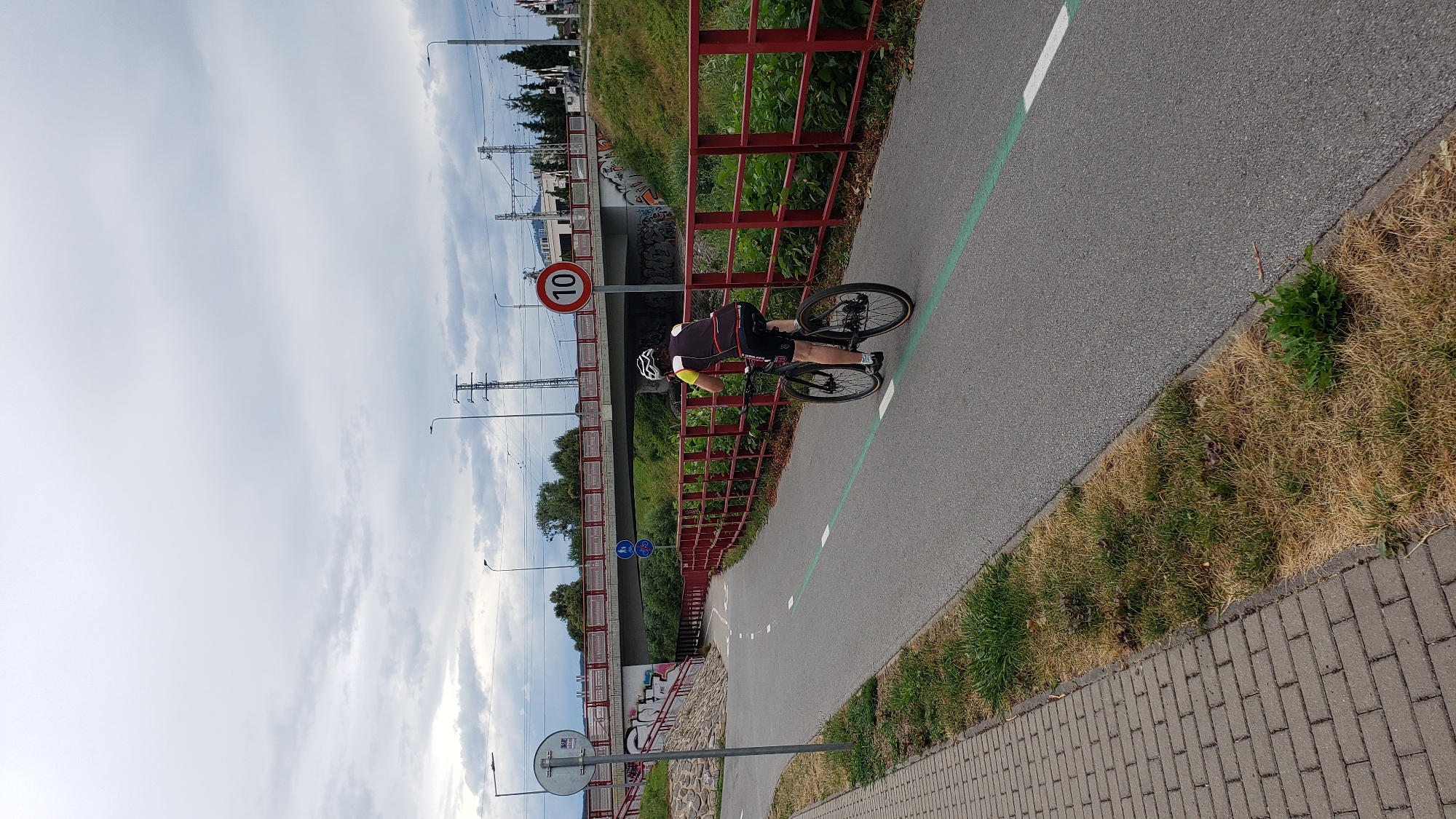 The image depicts a cyclist riding on a paved bike path in Prešov, Slovakia. The path is bordered by a red railing on the right and a cobblestone sidewalk on the left. A speed limit sign indicating "10" is visible on the right side of the path. In the background, a pedestrian bridge spans over a green area, and further in the distance, train tracks and power lines are visible under a cloudy sky. The cyclist, wearing a helmet and athletic attire, is heading away from the camera.