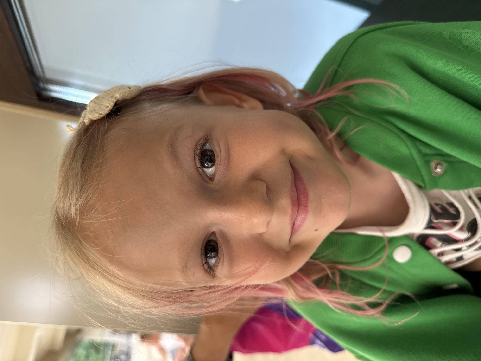 A close-up, head-and-shoulders shot shows a young Caucasian girl with blonde hair, featuring pink streaks, smiling and looking directly at the camera. She is wearing a green jacket over a white t-shirt with a graphic design. A small, cream-colored bow is visible in her hair. The background is out of focus, but suggests an indoor or outdoor setting with a light-colored wall and a hint of greenery. The lighting is natural and bright, illuminating the girl's face. There are no discernible landmarks or indicators of a specific city or location.
