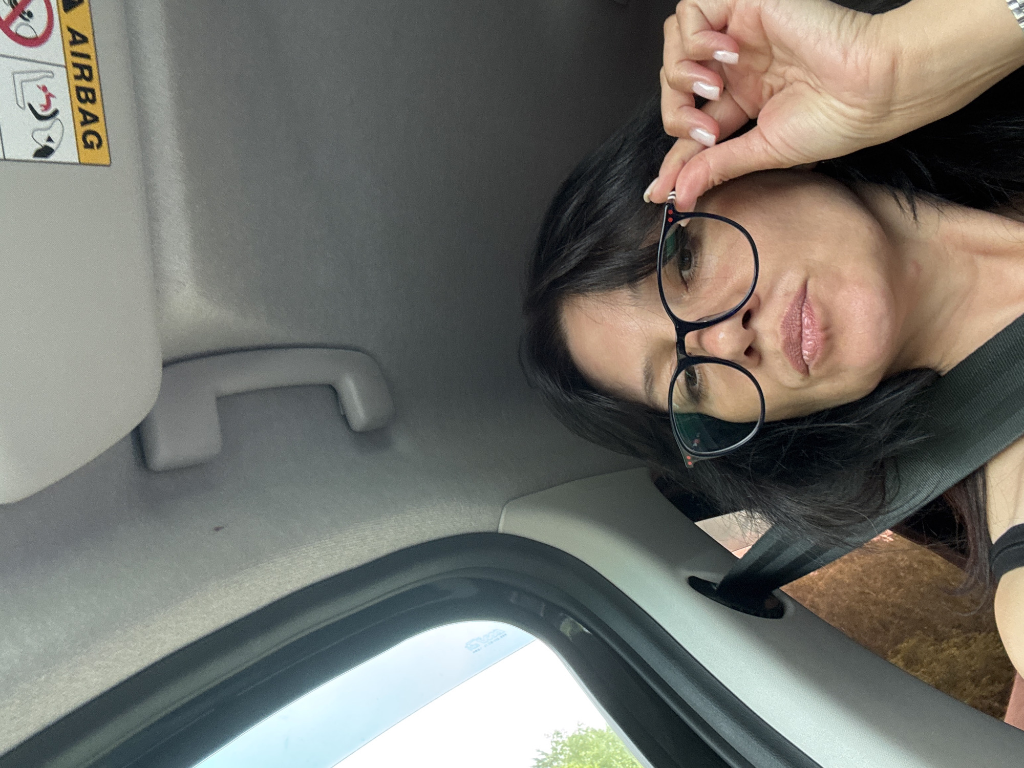 The image shows a woman in a car, adjusting her glasses. She has dark hair and is wearing a black tank top and glasses. The car interior is visible, including the headliner, a sun visor with an airbag warning label, and a seatbelt. The view outside the car window shows trees and sky. The provided location, Kazaevo, Russia, is not discernible from the visual information in the image.