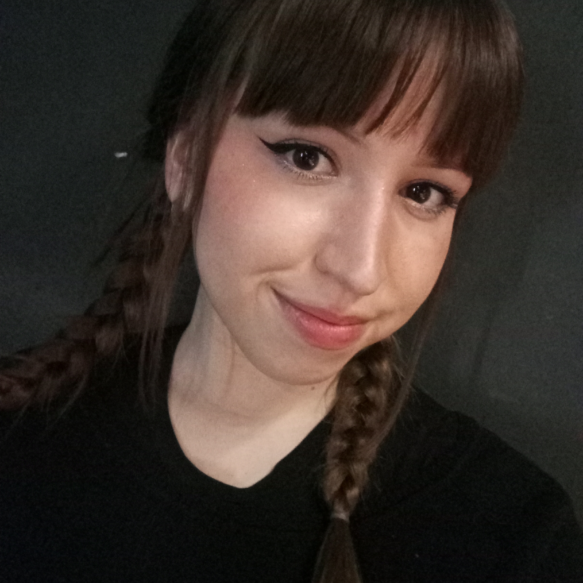 A young woman with brown hair and bangs, styled in pigtail braids, smiles at the camera. She is wearing a black shirt. Her eyes are lined with eyeliner. The background is a dark, solid color. There is no indication of this image being taken in the city of Chaikovsky, Russia.