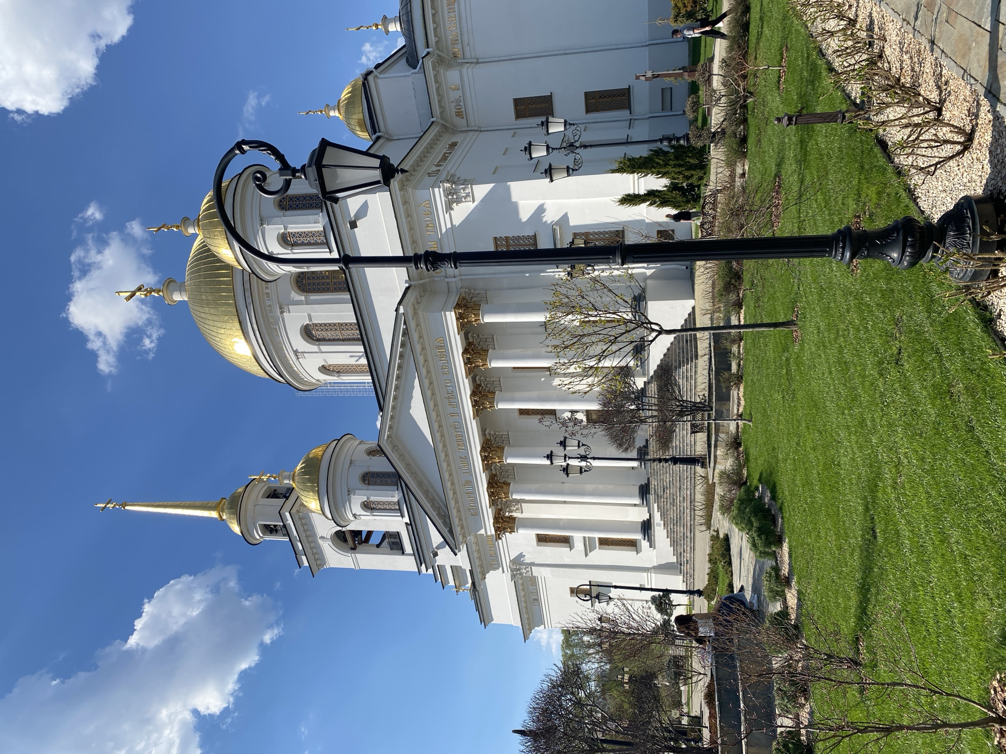 The image shows a white, ornate building with multiple golden domes, presumably a church, against a blue sky with scattered clouds. A black lamppost stands prominently in the foreground. A green lawn surrounds the building. There is a stone staircase in front of the building. Bare tree branches are visible near the lawn, suggesting early spring or late autumn. People are present but not easily discernible, given the angle and distance. The location is likely in Yekaterinburg, Russia.
