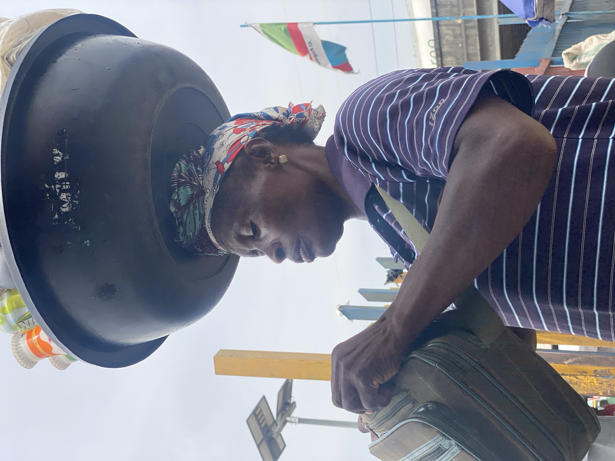 An outdoor, eye-level shot, likely taken on a bright day in an urban or market setting in Lagos, Nigeria. The main subject is an individual with dark skin, positioned slightly to the right of the frame, looking towards the left. They are wearing a dark blue or purple polo shirt with thin, light blue vertical stripes, and the brand "IZOD" is visible on the chest. A colorful headscarf, featuring red, blue, and white patterns, is tied around their head. A gold-colored stud earring is visible in their right ear.
The person is carrying a large, black, round basin or pot on their head, a traditional method of transporting goods. To the left of the basin, several plastic bottles with labels (one partially showing "TRK") are visible, possibly attached to or inside the basin. The person is also holding a light brown or beige shoulder bag or small suitcase with prominent zippers in their right hand, which rests near their hip. Their facial expression appears neutral and focused, with slightly parted lips.
In the background, a bright, overcast sky dominates. A multi-colored flag, primarily featuring white, green, and red horizontal stripes with some blue elements and illegible text, flies against the sky. A yellow horizontal beam or structure is visible in the mid-ground, along with what appears to be a solar panel street light in the lower background. Other indistinct structures are also present, hinting at an active environment.