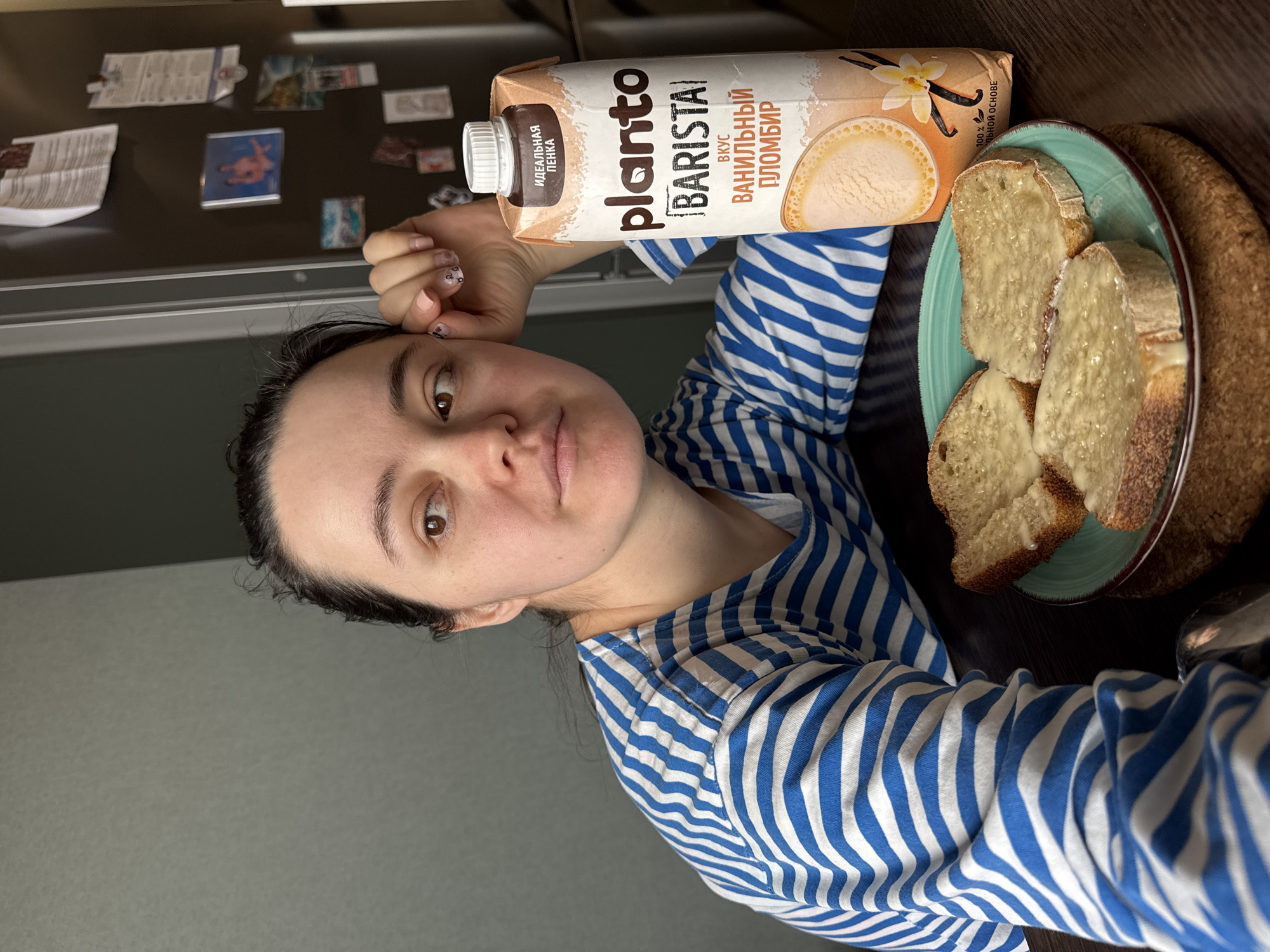 A woman is taking a selfie indoors, holding a carton of Planto Barista vanilla-flavored plant-based milk. She's wearing a blue and white striped shirt. To her right is a plate of bread slices spread with butter. The background shows a gray refrigerator with magnets on it and a dark wall. The product packaging is in Russian, indicating the image may be taken in a Russian-speaking region. The city of Izhevsk, Russia, is not discernible from the image.