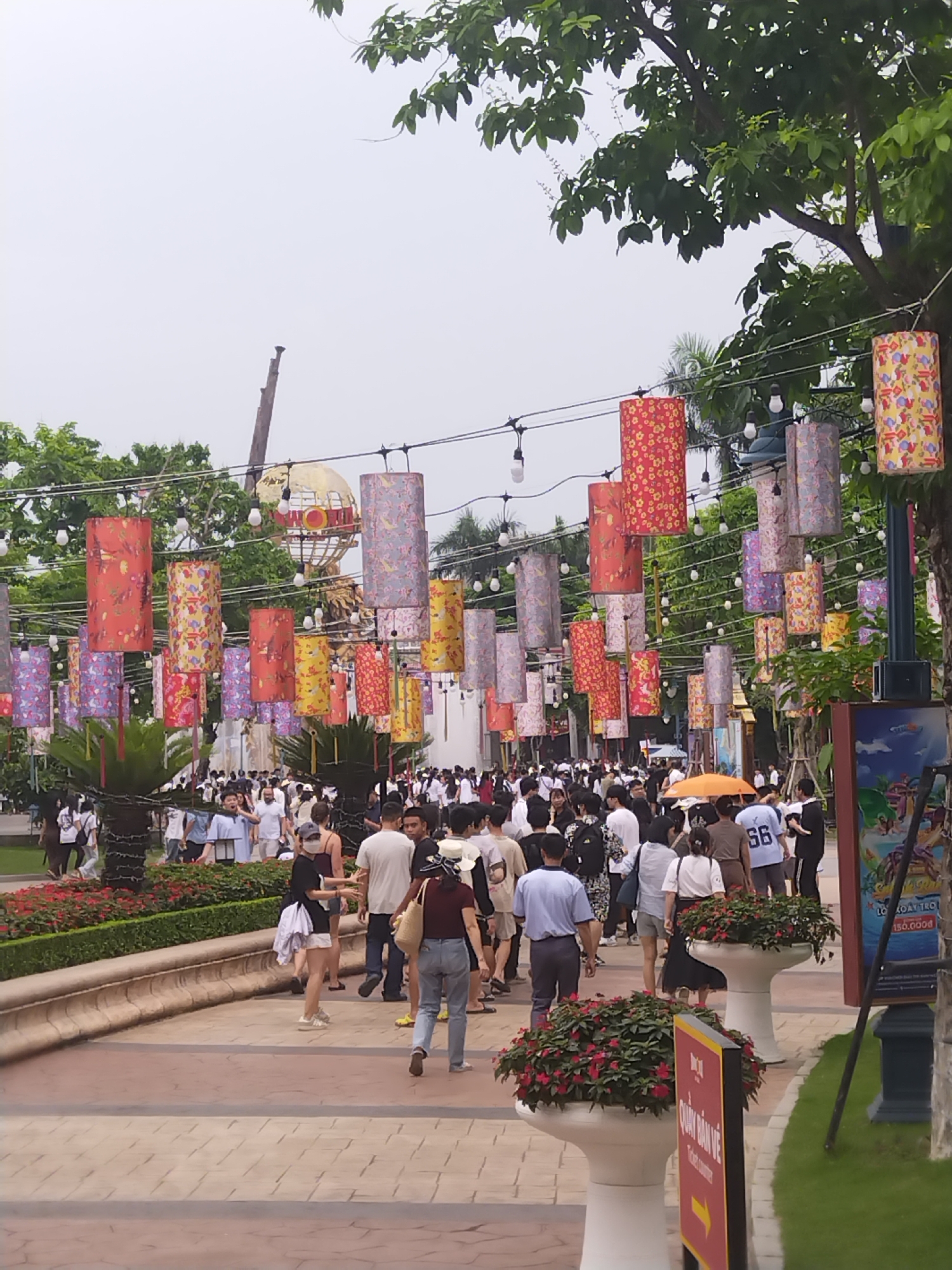 The image captures a bustling public space in Bai Chay Ward, Vietnam. A brick walkway extends into the scene, lined with manicured flowerbeds in white planters. Overhead, strings of lights and colorful cylindrical lanterns hang above a dense crowd of people. Individuals are walking in both directions along the path. Many appear to be tourists, wearing a variety of casual clothing. In the background, a tall structure resembling a globe on a pedestal is visible. Signs are on display near the right of the frame. The sky is overcast, casting a soft light across the scene.