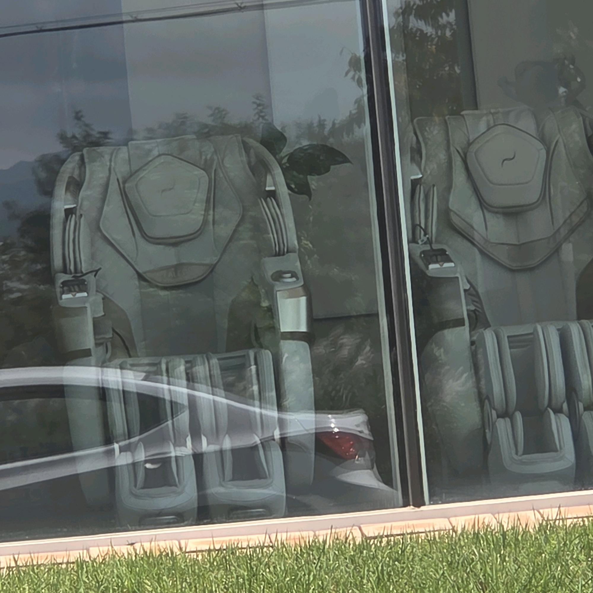 The image shows two massage chairs visible through a large window. The chairs are light grey and appear to be futuristic in design, with large padded sections for the back and legs. There are visible control panels on the side of the left chair. Reflections in the window show greenery and trees, suggesting an outdoor environment. The chairs are positioned indoors, possibly in a showroom or a private residence. The foreground shows a strip of green grass. The image appears to be taken in Gangneung-si, South Korea, but no specific landmarks or street views are visible.