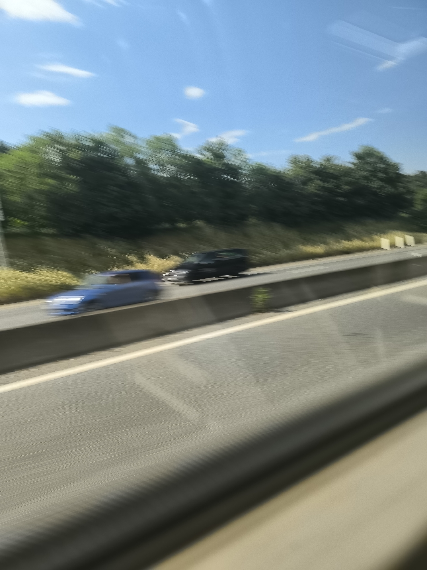 The image shows a highway near Saint-Chamond, France. There are two cars traveling on the highway, one blue and one black. The image is blurred, suggesting it was taken from a moving vehicle. There are trees and foliage visible in the background, under a bright sky with scattered clouds. The road surface is asphalt and has a concrete barrier.