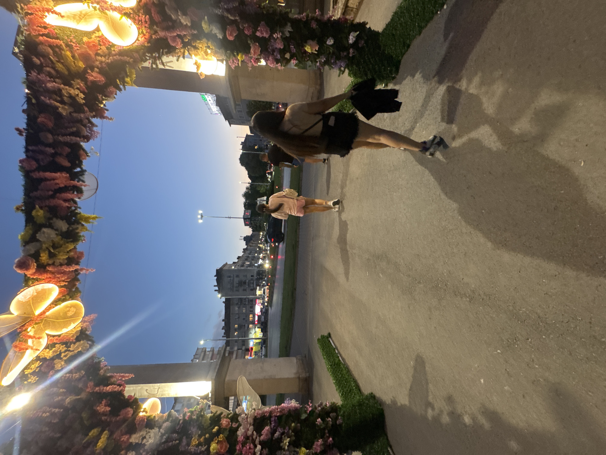 The image shows an outdoor evening scene in Shumen, Bulgaria. Two female figures are walking on a paved sidewalk adjacent to a street. The closest figure is wearing a dark top and shorts, with a dark jacket hanging from one arm. The other figure is wearing lighter-colored clothing, possibly a pink dress. Streetlights illuminate the scene and building facades. A decorated archway frames the image, adorned with flowers and illuminated butterfly-shaped lights. The camera angle is tilted to the side.