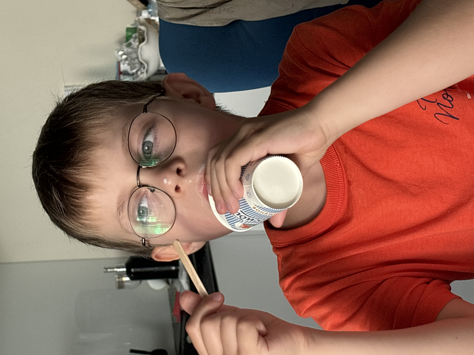 A young boy with glasses is shown from the chest up, eating from a small cup with a wooden stick. He is wearing an orange t-shirt. There is a bit of white residue around his mouth, suggesting he has been eating something creamy like yogurt or ice cream. The background is blurred, but it appears to be an indoor setting with a grey wall on the left and a blue chair on the right. The boy's eyes are blue and he is looking to his left. The image quality is good, and the lighting is natural.