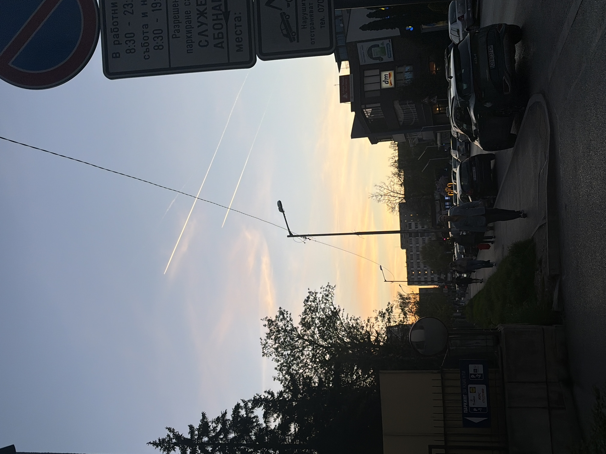 The image depicts an urban scene in Sofia, Bulgaria, at twilight. The sky is streaked with contrails from aircraft. Street signs are visible in Bulgarian; one indicates parking restrictions. A "no parking" sign is also visible. Buildings line the street, and parked cars are aligned along the curb. People are walking along the sidewalk. A KFC parking sign can be seen. A DM drugstore sign is also seen. Car registration CO 8737 CH can be seen.