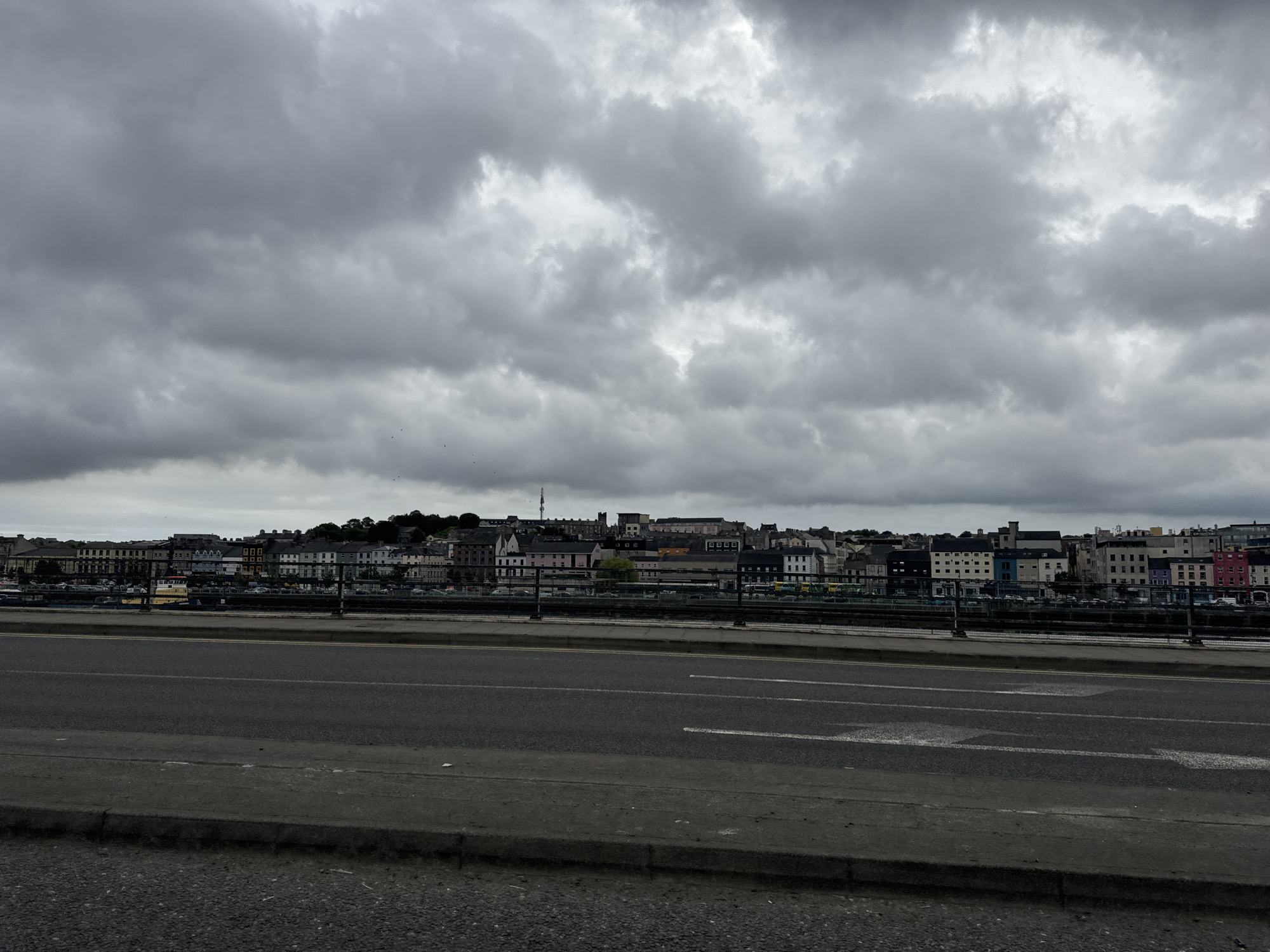 The image shows a wide shot of a city skyline across a body of water, with a road in the foreground. The sky is overcast with heavy, grey clouds, suggesting inclement weather. The city itself features a dense collection of buildings of varying heights and colors, including multi-story structures and some with distinctly colored facades like pink, blue, and yellow. A prominent yellow boat is visible on the water near the shore, in front of the buildings. A road with visible lane markings runs parallel to the city skyline, with a concrete barrier separating it from the water. The foreground consists of the road and a concrete curb, with patches of gravel and debris on the ground. The overall impression is one of an urban waterfront scene on a cloudy day in Ireland.