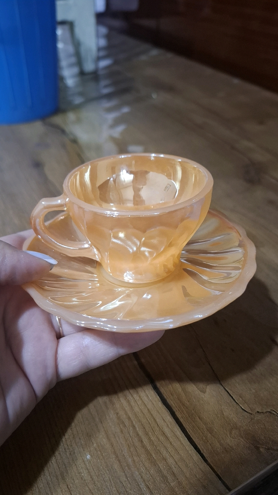 A close-up, eye-level shot shows a hand holding an iridescent orange glass teacup and saucer. The teacup is octagonal with a fluted design and a delicate handle, while the saucer is similarly fluted and scalloped around the edge. The teacup and saucer are made of glass with a pearlescent sheen that reflects light and creates a rainbow effect. The background consists of a wooden table with visible grain and a blurred blue object. The scene is devoid of any Shymkent-specific landmarks or context.