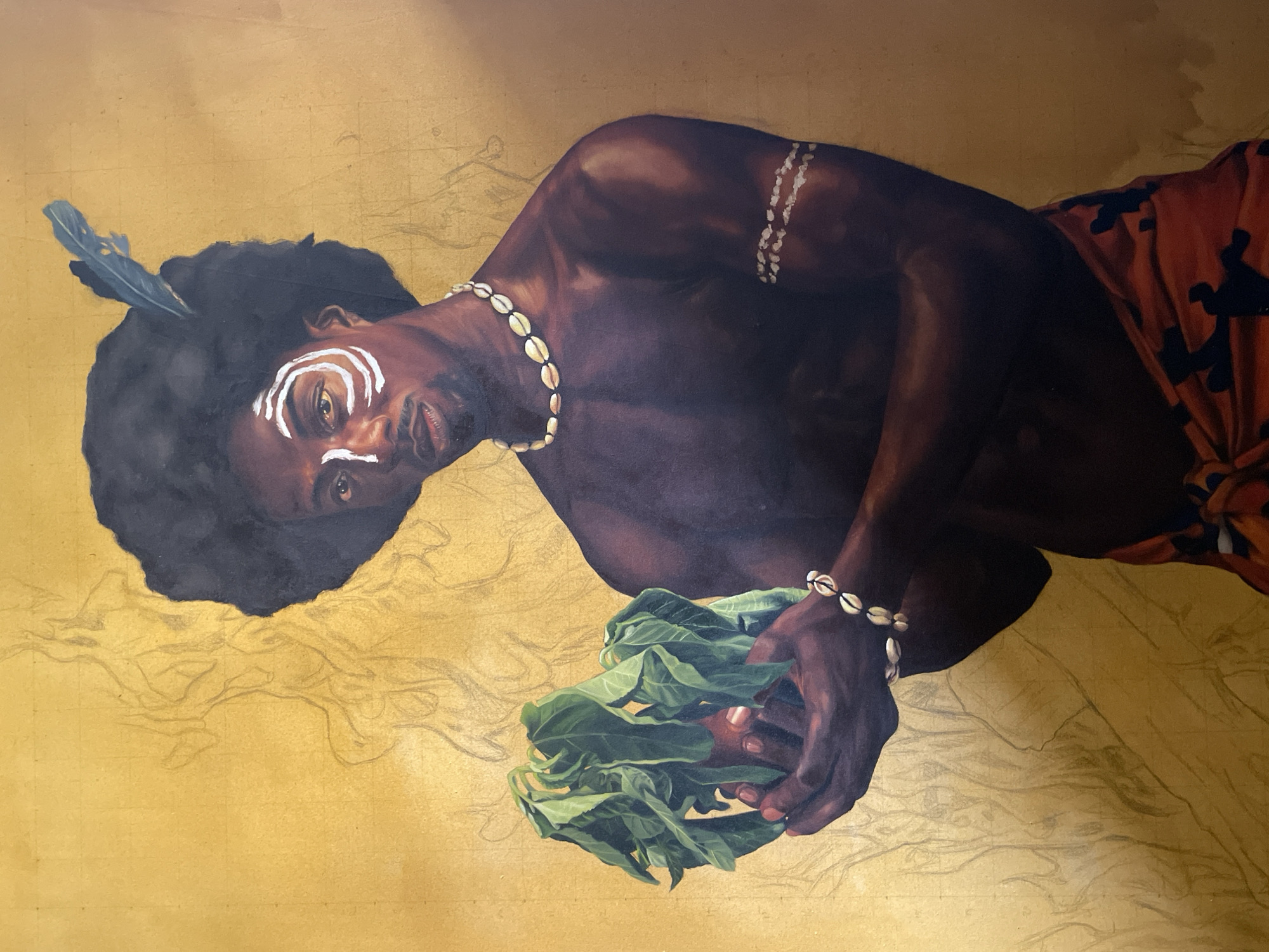 A painting depicts a dark-skinned man with black curly hair. He wears a blue feather in his hair, white face paint markings, and a seashell necklace and bracelet. He is holding green leaves in his hands. He wears a red and orange patterned cloth wrapped around his lower body. The background is gold with faint pencil sketch lines. The image does not depict the city of Ado, Nigeria.