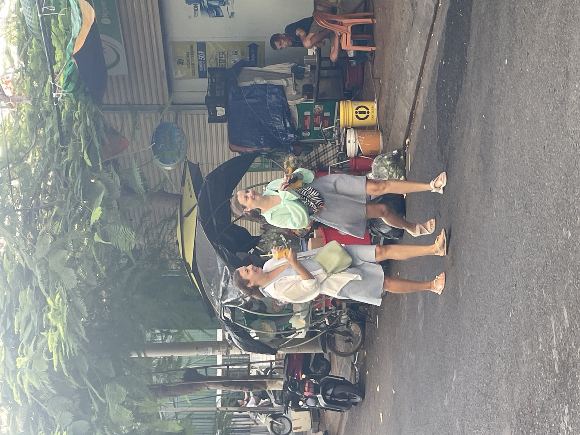 Two women are walking on a paved street in front of a building with a corrugated metal roll-up door. The woman on the left, wearing a light blue dress and a white open shirt, is carrying a light green purse over her shoulder and a clear cup with an orange beverage in her right hand. The woman on the right, wearing a light green top and a gray skirt, is also holding a clear cup with an orange beverage and has a black and white patterned bag slung across her chest. Behind them, a street vendor's cart with a black canopy is visible, filled with what appear to be flowers and other items. A motorcycle is parked to the left of the cart. In the background, a man is sitting at a table with a chair, and other objects like buckets and tarps are scattered around. The scene is set under a canopy of lush green trees, suggesting an outdoor, possibly commercial, area in Bac Nha Trang Ward, Vietnam.