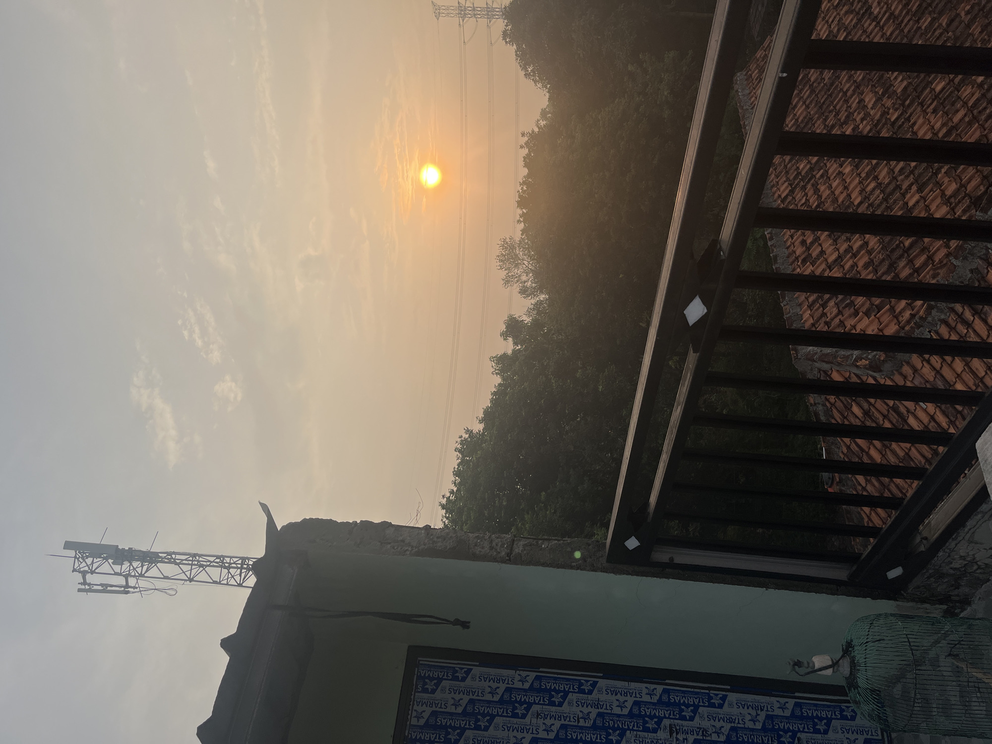 In Pasir Gadung, Indonesia, a scene unfolds at sunrise, with the sun a bright orb low in the hazy sky, casting a warm, golden light. The sky is partly cloudy, with wispy formations scattering the light. In the foreground, the edge of a light green building is visible, with its roofline and a partially visible wall. A metal railing with dark vertical bars runs along the top of this building, overlooking a densely forested area. Beyond the trees, power lines stretch across the landscape, supported by tall transmission towers, indicating the presence of infrastructure within or near the city. The roof of another building, covered in red terracotta tiles, is visible beneath the railing. To the left of the frame, a green birdcage sits on a ledge.