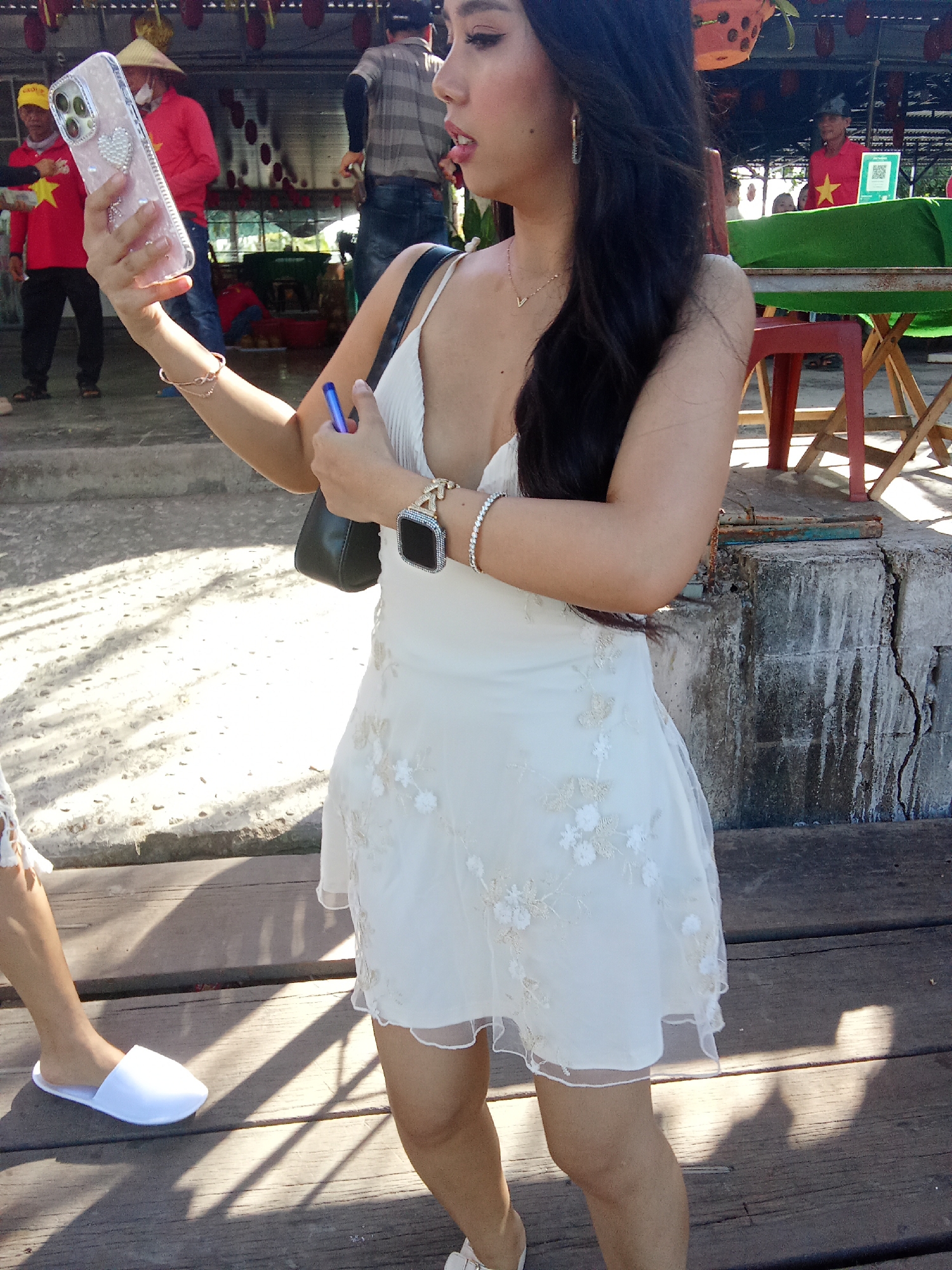 A woman is standing on a wooden boardwalk, holding a phone and a pen. She is wearing a white dress with floral embroidery, a silver bracelet, and a watch on her left wrist. Her hair is long and dark, and she has prominent makeup. Beside her, another person's legs and feet are visible, wearing white slippers. In the background, several people are present in what appears to be an outdoor market or food stall area. One man wears a conical hat and a red shirt with a yellow star, and another wears a striped shirt and jeans. There are also tables covered with green fabric, chairs, and stalls with hanging decorations. The scene is brightly lit, suggesting daytime. The presence of Vietnamese flags (yellow star on a red background) indicates the location is likely in Vietnam. Given the context of markets and the general ambiance, this scene could be situated in a riverside market area or a food street in Đà Nẵng, Vietnam, possibly near the Han River.