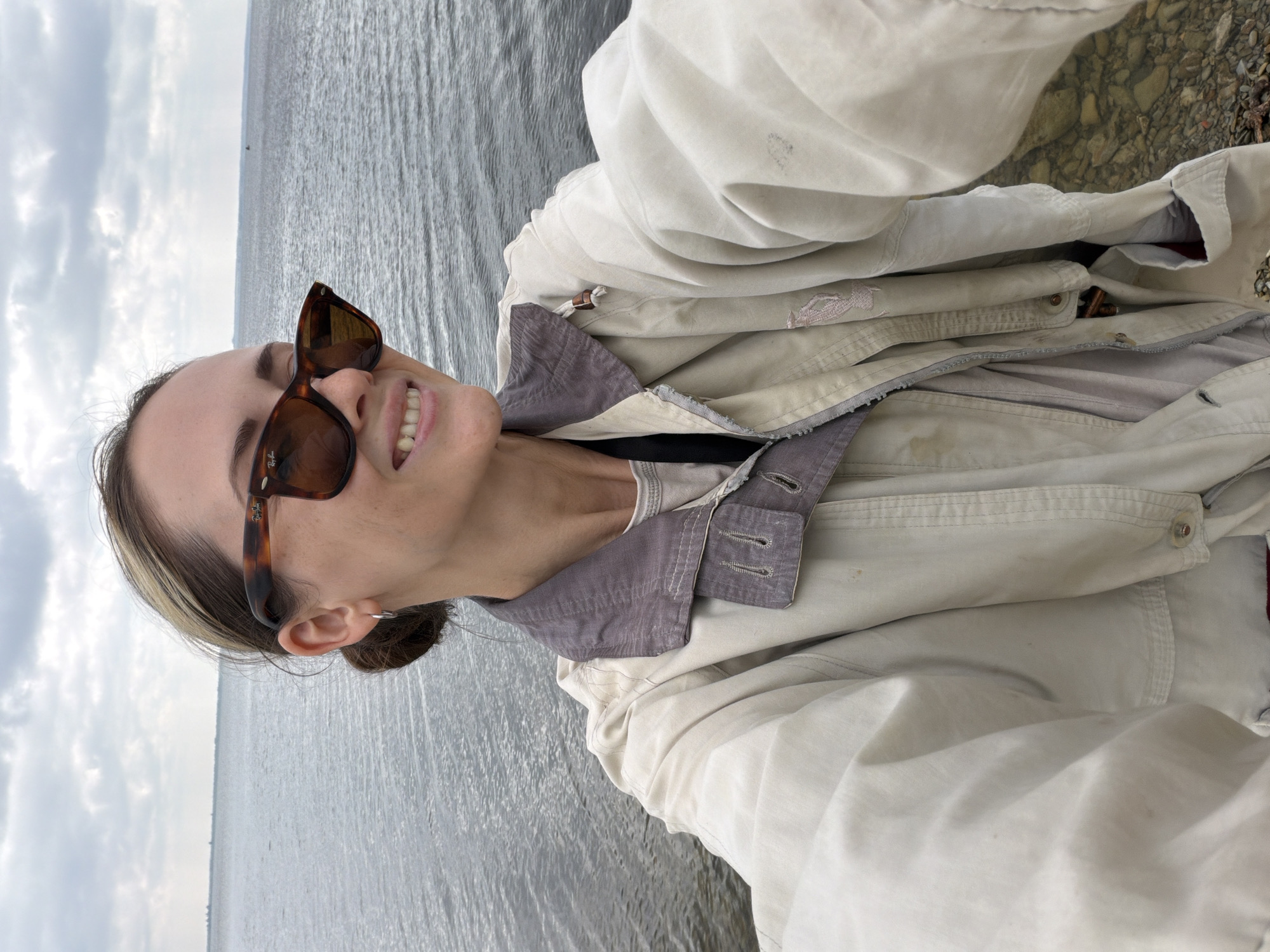 A woman is taking a selfie outdoors with a body of water in the background. She is wearing sunglasses, a light-colored jacket with a purple collar, and a light-colored t-shirt. The body of water appears to be a large river or lake. The sky is cloudy. The ground in the foreground consists of small rocks and pebbles. The woman's hair is pulled back into a bun. She is smiling. The location is the Volga River embankment in Kamyshin, Russia.