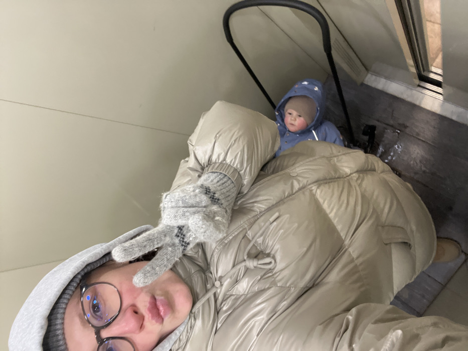A woman and an infant are depicted within a confined space, likely an elevator, in Khimki, Russia. The woman occupies the majority of the frame, angled downwards, wearing a shiny light beige puffer jacket, a grey hooded garment beneath, dark-framed glasses, and light grey knitted gloves with a black patterned design. Her right hand extends a "peace" sign, and her lips are pursed. Visible below her and to the right is an infant seated in a stroller, primarily identifiable by a black handlebar. The infant wears a blue hooded outfit with light markings and a light brown knitted cap, exhibiting rosy cheeks and gazing upwards. The immediate surroundings consist of plain light beige walls and a dark floor, possibly wet or reflective, transitioning to a lighter metallic threshold near what appears to be a doorframe.
