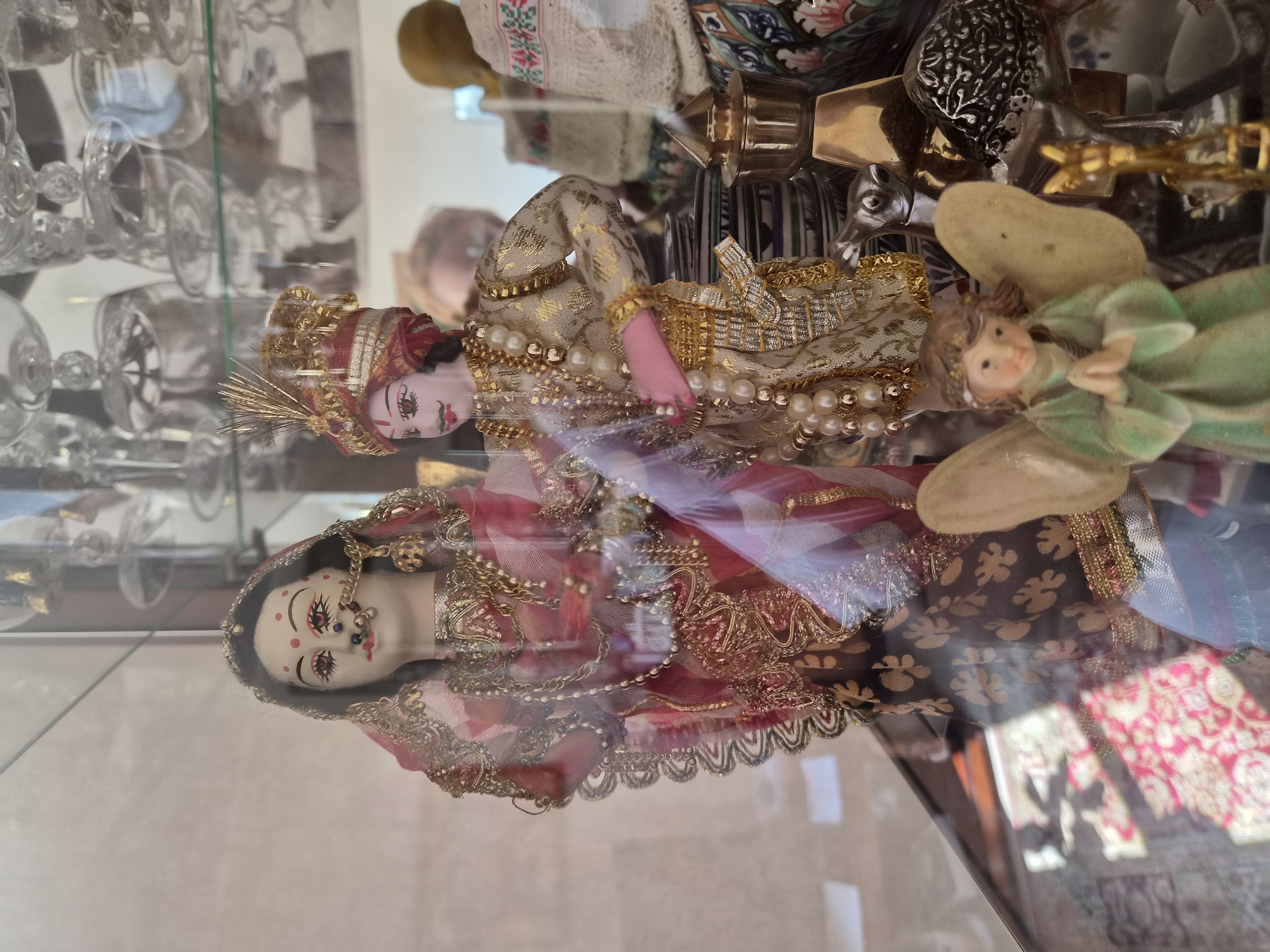 This is a close-up, eye-level photograph of a display case containing various figurines. The figurines are highly detailed, and appear to be traditional Indian dolls dressed in elaborate clothing. The doll on the left is a female figure wearing a red and gold sari, adorned with jewelry, including a nose ring and a headdress. The doll on the right is a male figure, also dressed in rich, ornate attire with a turban and pearls. In the foreground, a small angel figurine with green wings and a green robe is placed in front of the larger dolls, with its hands clasped in prayer. Behind the angel and to the right of the larger dolls are other decorative objects, including what appears to be a stack of ceramic plates and some metal decorative items. The display case itself is made of glass, and the background is blurred, showing reflections of other items, likely in a shop or museum setting. There are no discernible people actively engaged in interactions within the frame. The lighting is soft, and the overall impression is one of cultural display and artistic representation. The location is identified as Yelabuga, Russia.