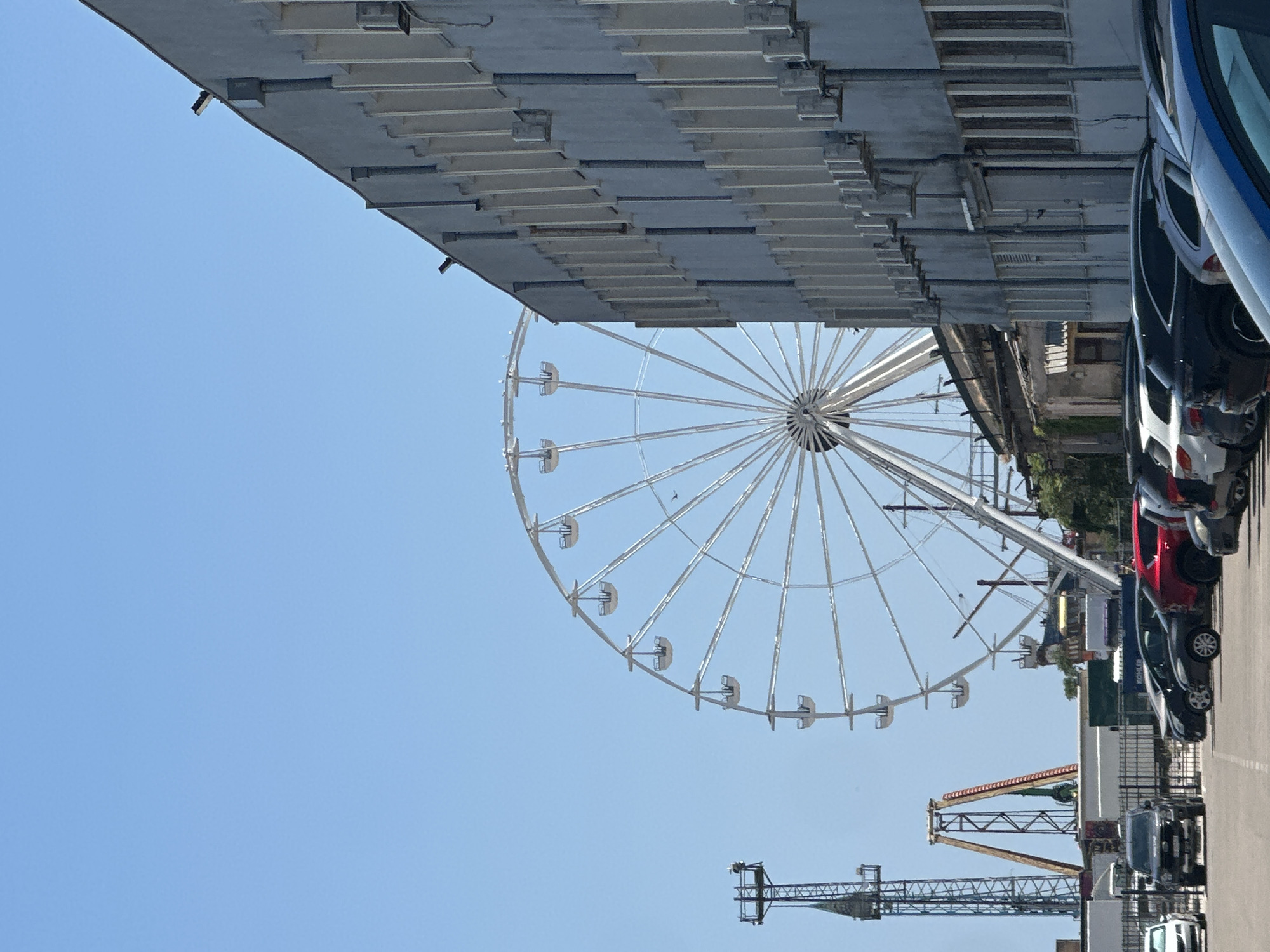 A Ferris wheel stands tall against a clear blue sky in this outdoor scene. In the foreground, a gray, multi-story building with numerous windows and air conditioning units lines the right side of the image. A parking lot filled with various cars, including a black SUV, a red car, and several other vehicles, is visible in front of the building. To the left of the Ferris wheel, a tall, slender tower with a platform at the top is present, and further to the left, a ride with a suspended structure can be seen. The Ferris wheel itself is large and white, with many enclosed gondolas suspended from its rim. The overall impression is that of an amusement park or fairground area, likely on a bright, sunny day.