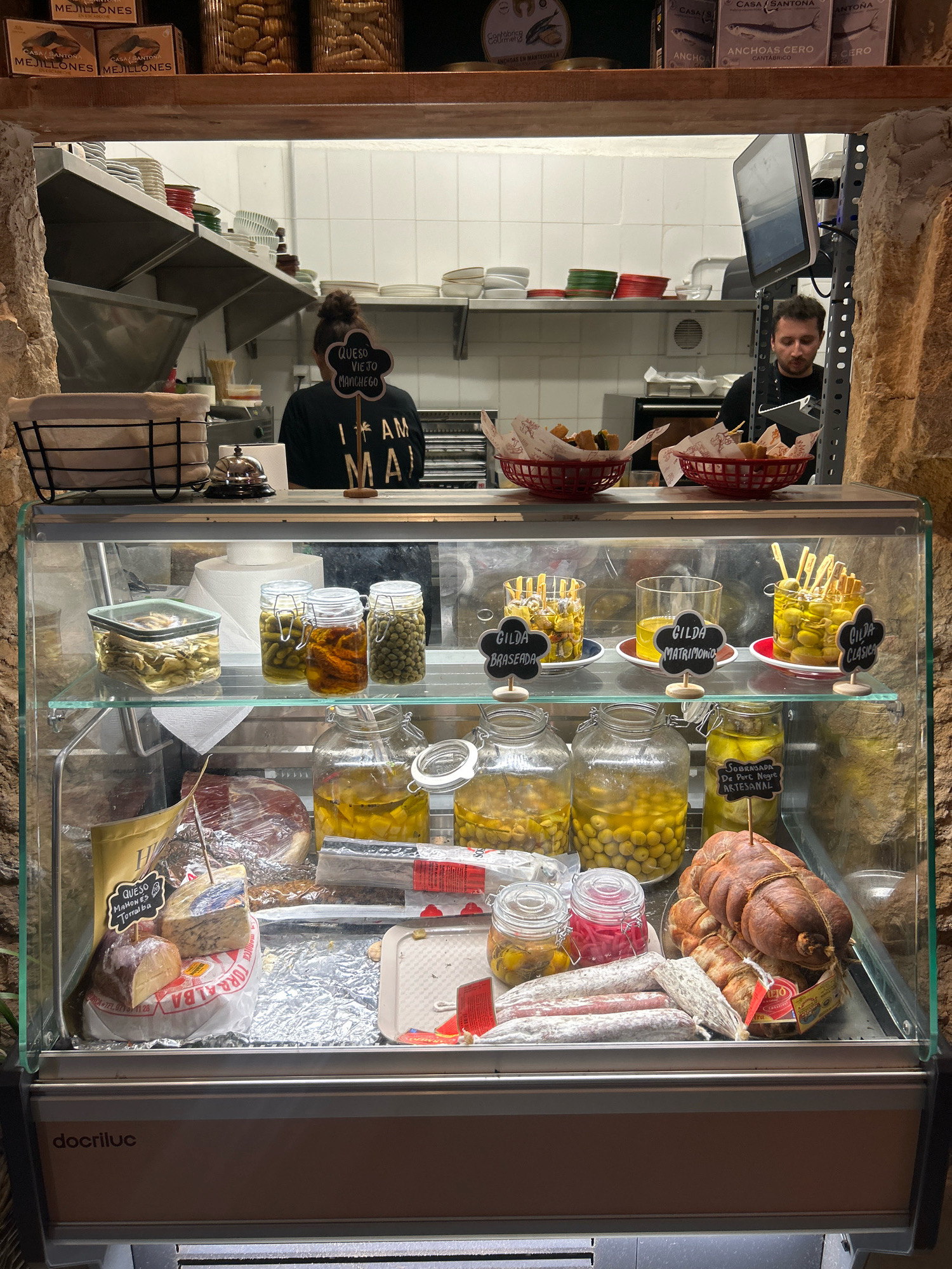 The image shows a food display case in a deli or restaurant in Palma de Mallorca, Spain. In the background, two people are visible working in what appears to be a kitchen area. One person, wearing a black t-shirt with "I AM MAJ" written on it, stands behind a counter. Another person, a man wearing a black shirt, is looking at a tablet.
The display case is filled with various food items. On the top shelf, there are jars of olives, sun-dried tomatoes, and what appear to be pickled vegetables. There are also small glasses containing olives with skewers. Several black chalk signs are present, some labeling the items as "GILDA BRASERA," "GILDA MATRIMONIO," and "GILDA CLASICA."
The lower shelf of the display case contains more food items, including a large piece of cured meat, several types of salami or cured sausages, a wheel of cheese labeled "QUESO MAHONES TORRALBA," and a smaller wedge of cheese. There are also jars of olives and other preserved items. A roll of aluminum foil is visible in the lower left corner.
The overall environment appears to be a commercial food establishment, with shelves lined with various food products and packaging in the background. The display case itself is made of glass and metal, with internal lighting. The bottom of the display case has a branded panel that reads "docriluc." The exterior of the establishment has a stone-like facade, suggesting an older building or a design choice to evoke a rustic feel.