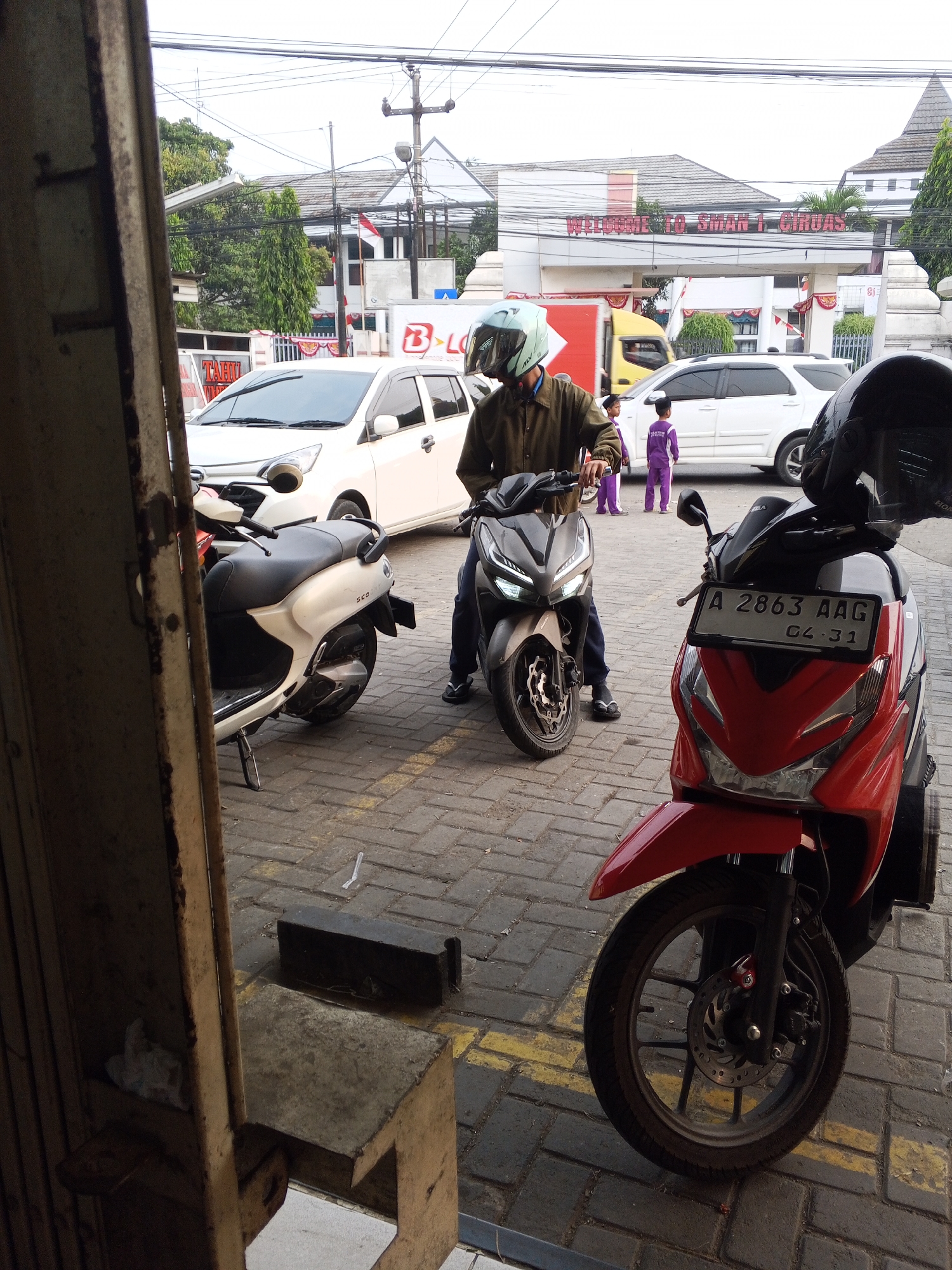 The image depicts a street scene in Ciruas, Indonesia, with a prominent sign reading "WELCOME TO SMAN 1 CIRUAS." In the foreground, two motorcycles are parked on a brick-paved area. The motorcycle on the right is red and black, with its license plate showing "A 2863 AAG 04-31." The motorcycle on the left is gray, and a person wearing a brown jacket and a light blue helmet is seated on it, holding the handlebars. Behind the motorcycles, a white car is visible. Further back, two children in purple uniforms are standing near a white SUV. The background features buildings with tiled roofs and trees, suggesting an urban or semi-urban environment. The overall atmosphere is that of a typical day on a street near a school.