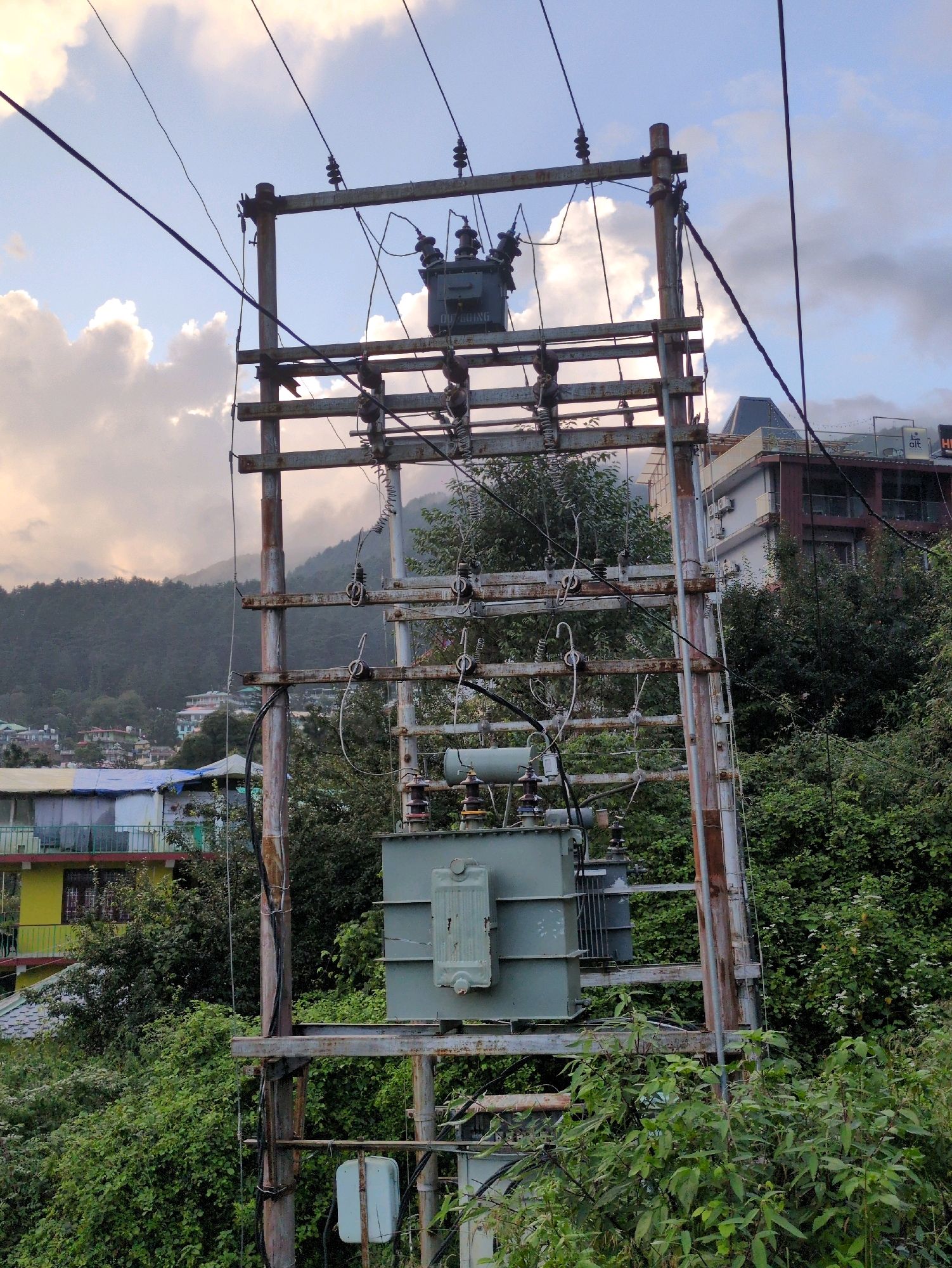 The image depicts an outdoor scene in Haini, India, focusing on an electrical transformer and pole assembly. The transformer, a large, light green metal box, is mounted on a rustic, rusty metal pole structure. Wires extend from the transformer upwards and are supported by insulators attached to horizontal beams on the pole. Above the main transformer, a smaller, darker transformer unit is visible. The pole structure itself is showing signs of corrosion and weathering.
The surrounding environment is lush and green, with dense foliage and trees obscuring much of the background. In the distance, buildings are visible on a hillside, suggesting a semi-urban or town setting. The sky above is partly cloudy, with dramatic cloud formations and a soft, diffused light, indicative of either early morning or late afternoon. No people are present in the image. The overall impression is one of utilitarian infrastructure set against a natural, hilly landscape.