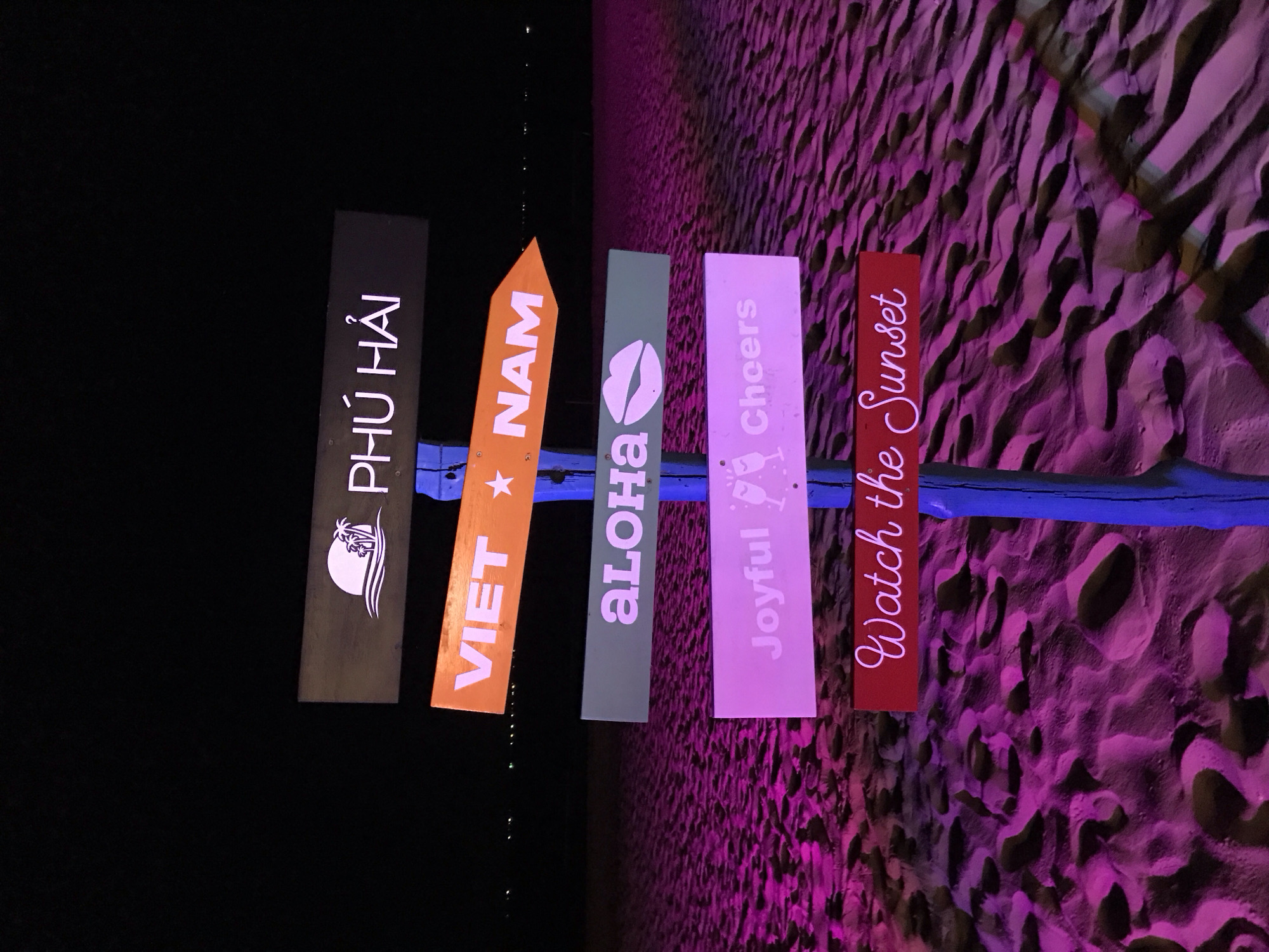 This media file shows a series of signs hanging on a wooden plank, set against a backdrop of sand dunes illuminated by purple light. The signs are arranged vertically and appear to be made of wood. From left to right, the signs read: "PHÚ HẢI" with a silhouette of palm trees and a beach scene, "VIET NAM" with a star, "ALOHA" with a kiss mark, "Joyful Cheers" with a champagne glass, and "Watch the Sunset". The sand dunes have a textured appearance, suggesting a beach or desert landscape. The lighting is dramatic, casting deep shadows and highlighting the contours of the sand, creating an evening or nighttime atmosphere. The overall mood is festive and inviting, likely indicating a celebratory event or a destination marker. The presence of Vietnamese text suggests a location in Vietnam, specifically Phu Thuy Ward, as indicated by the provided location context. There are no people visible in the media file.