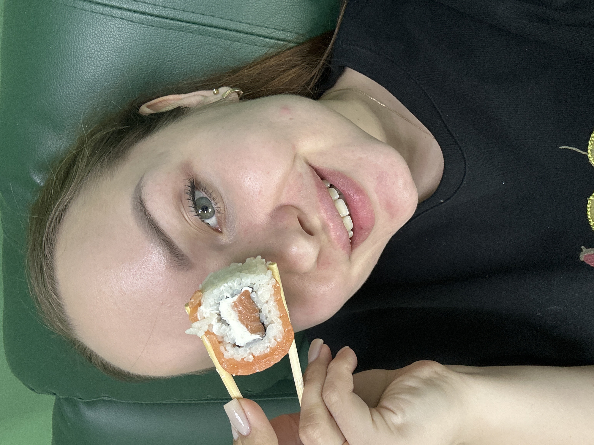 A woman is holding a sushi roll with chopsticks in front of her face. She is lying down on a green leather-like surface. The woman has fair skin, light brown hair, and blue eyes. She is wearing a black t-shirt and a gold necklace. Her nails are painted light pink.
I'm unable to identify the location as Chaikovsky, Russia. There are no obvious landmarks or other identifying elements visible in the image to make this determination.