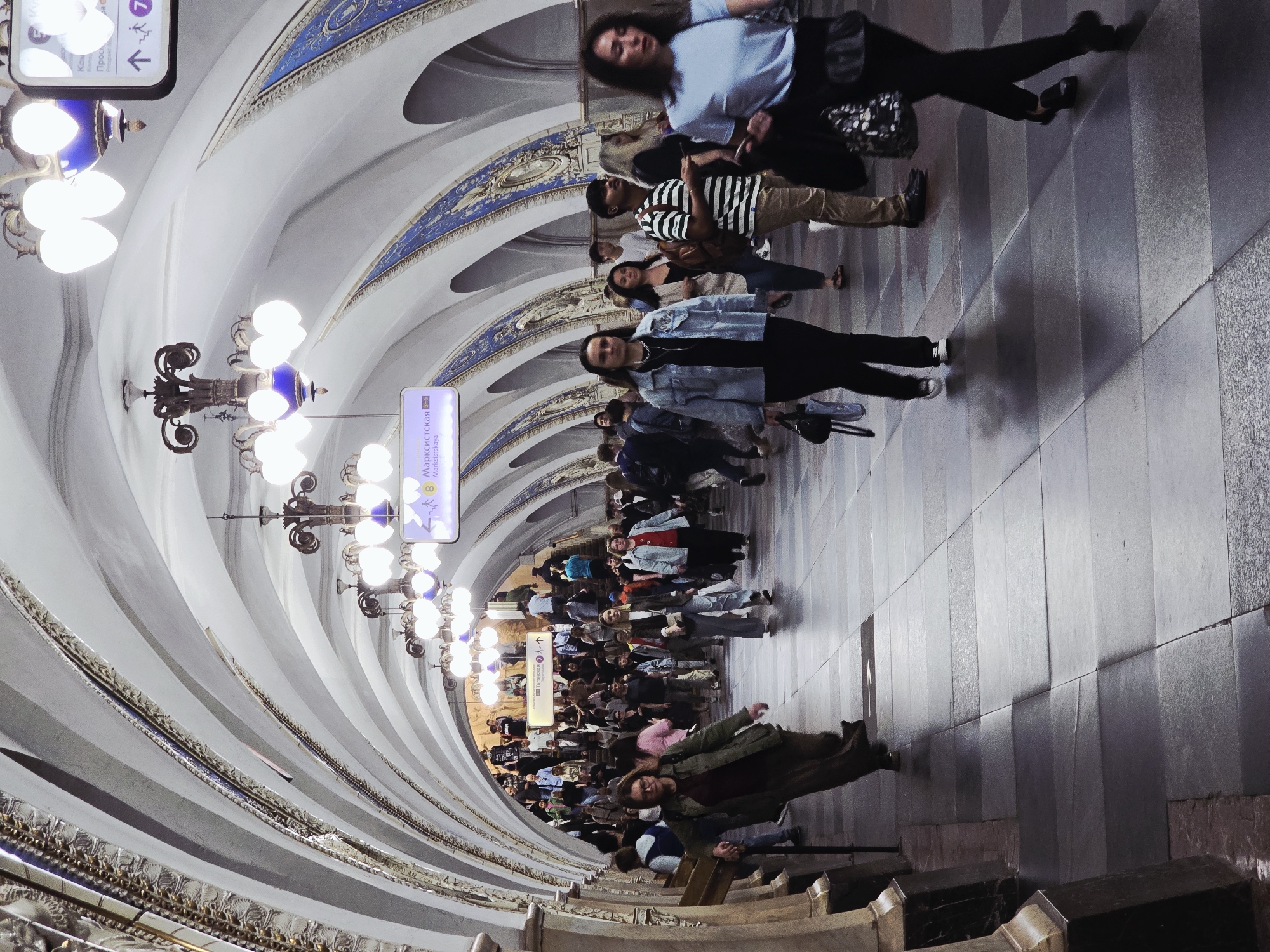 The image depicts a crowded underground tunnel, likely a metro station, in an unknown city. The tunnel has a high, arched ceiling with decorative elements and rows of chandeliers. Numerous people are walking through the tunnel, some appearing to be in motion. The floor is tiled with a pattern. The tunnel is illuminated by the chandeliers and other light sources. Station signage is visible.
