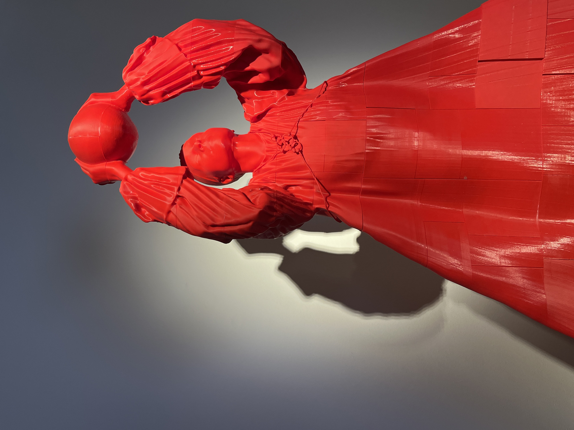 The image shows a red, glossy sculpture of a woman holding a ball. The sculpture is mounted on a gray wall. The woman is wearing a long dress or robe. The sculpture is likely located in Las Palmas, Spain.