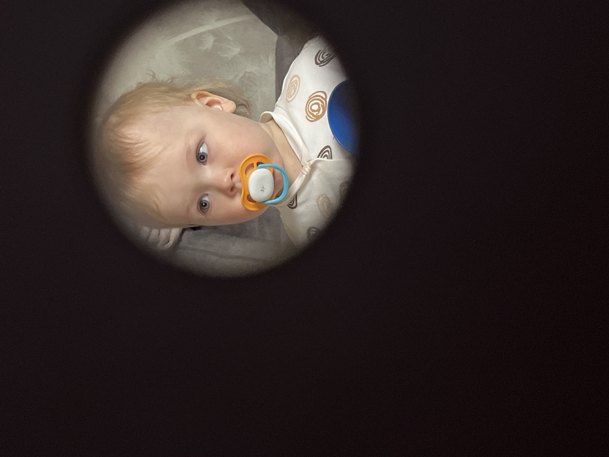 A young child with blond hair and blue eyes is looking at the camera with a pacifier in their mouth. They are wearing a light-colored shirt with circular patterns. The child is seen through a circular opening, with the surrounding area being dark and out of focus. The setting appears to be indoors. There is no indication of the location being in Chaikovsky, Russia, or any other specific place.