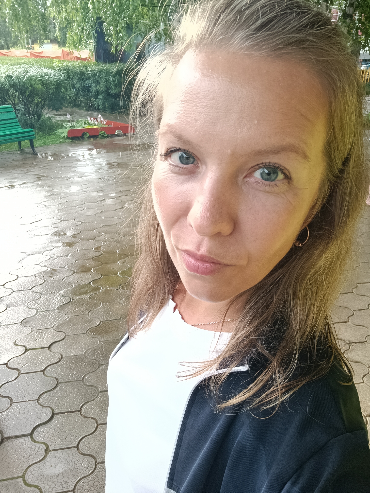 A woman with blonde hair and blue eyes is taking a selfie outdoors. She is wearing a white top and a dark jacket. The ground is wet and appears to be paved with interlocking stones. In the background, there are trees, bushes, and a green bench. There are also some red planters with flowers and orange tarps. The overall scene suggests a park or outdoor public space in Chaikovsky, Russia, possibly after a rainfall.