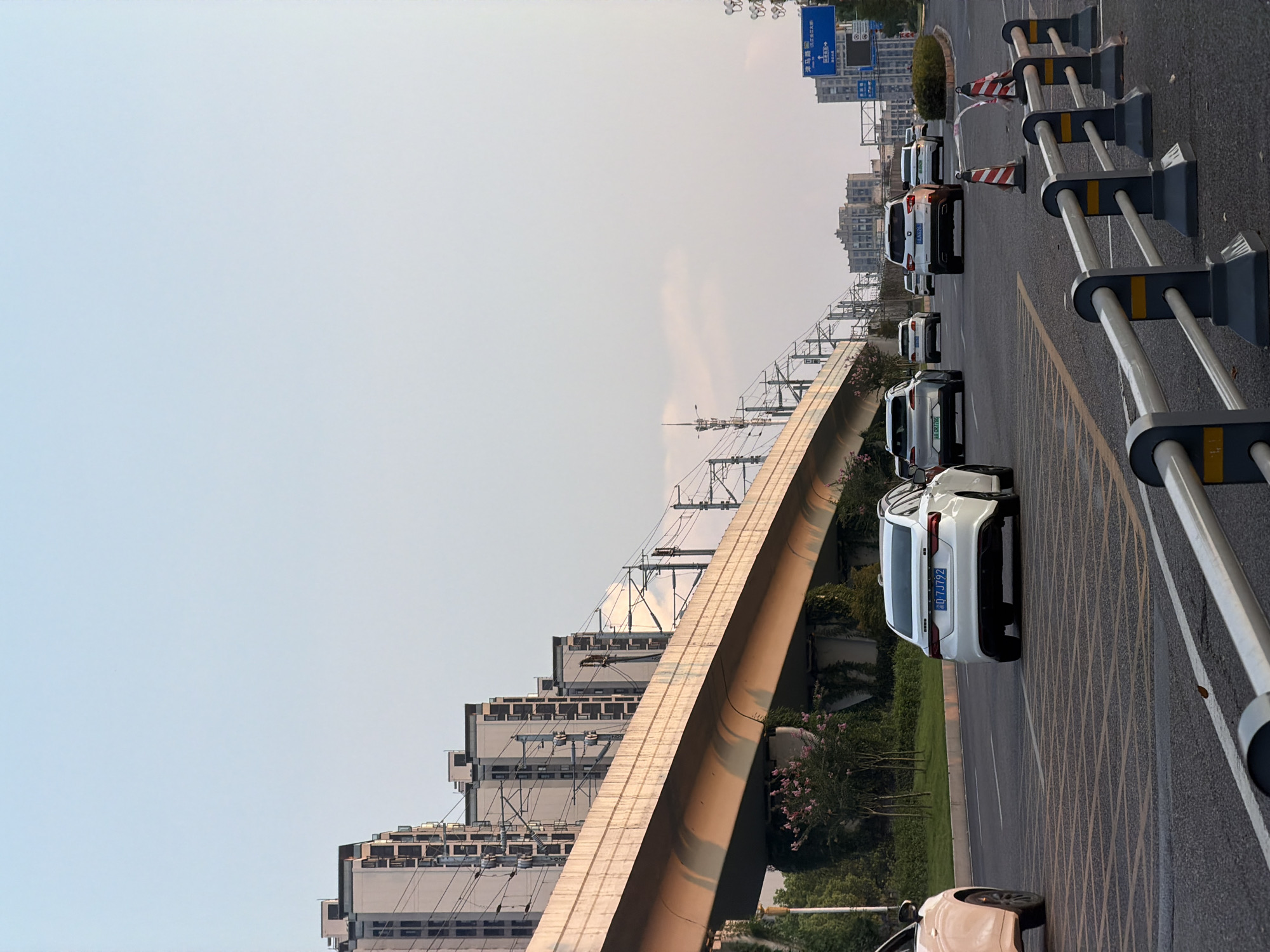 This is a vertical, eye-level, medium shot of a road in Jiangjin, China. The scene is illuminated by the warm glow of late afternoon sun. In the foreground, a silver sedan with license plate 0-DJ7J92 is driving away from the camera on the right side of the road. In the background, several other vehicles are also driving in the same direction. Above the road, an elevated train track stretches across the frame, supported by concrete pillars. Beyond the train track, a row of modern apartment buildings stands tall. The sky above is a pale blue, with a few wispy clouds. The road is marked with white and yellow lines, and traffic cones are visible on the right side of the road.