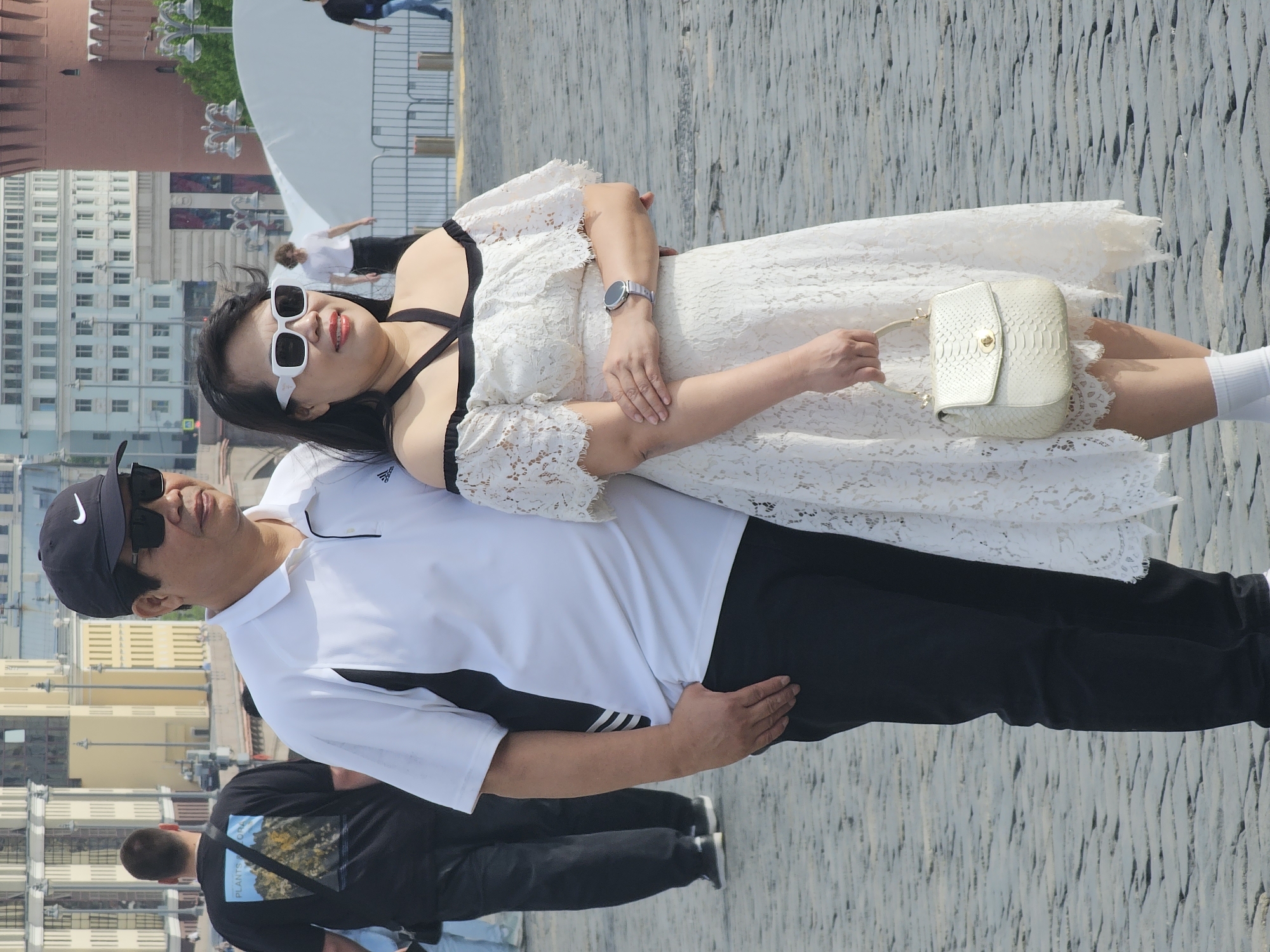 The image shows a couple posing outdoors. The woman is wearing a white lace dress with black straps and white sunglasses. She is holding a white textured handbag and wearing a watch. The man is wearing a white polo shirt with black accents, black pants, sunglasses, and a black baseball cap. They appear to be standing on a stone surface. In the background, there are buildings and architectural structures, including a red brick structure resembling a tower, typical of Moscow’s Red Square landmarks.