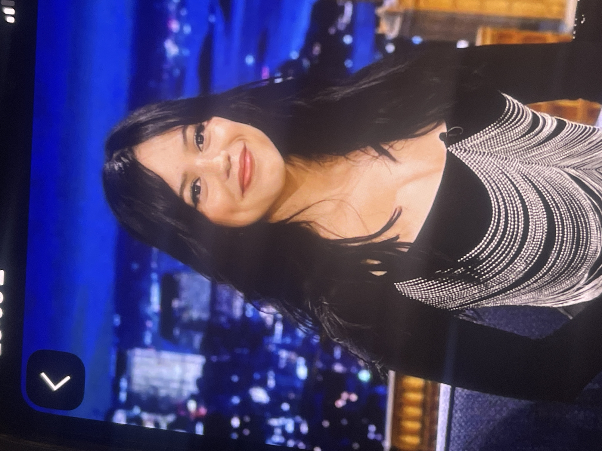 A young woman with long, dark hair and fair skin is captured in a vibrant image, smiling warmly directly at the viewer, with visible dimples. Her head is tilted, and her expression suggests cheerfulness and engagement. She is wearing a black long-sleeved garment, possibly a top or dress, which is intricately embellished with striking silver, beaded fringe details that drape over her shoulders and across her upper chest. These fringes create a dynamic, waterfall-like pattern.
The background is a deep, luminous blue, featuring blurred lights that give the impression of a stylized city skyline at night, or a backdrop within a television studio or stage setting. This suggests an indoor environment, likely for a public appearance, interview, or performance.
On the far left side of the image, a partial digital interface is visible, including a dark rounded square button with a white "V"-shaped arrow, indicating an interactive element from a screen, such as a video player or navigation control. The overall media file appears to be a photograph of a screen, capturing a moment from a broadcast or recorded event.