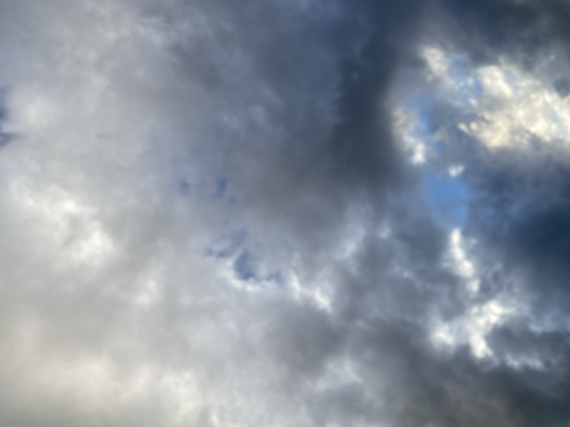 The image shows a cloudy sky with patches of blue visible. The clouds are dark and varied in texture, suggesting they may be a mix of nimbostratus and cumulus clouds. Sunlight filters through some of the clouds, creating brighter areas and highlighting the depth and movement of the cloud formations. The perspective is from below, looking upwards. The image is slightly out of focus, giving it a soft, atmospheric quality. The scene is outdoors and appears to be a natural sky formation. No human subjects or specific city landmarks are identifiable.