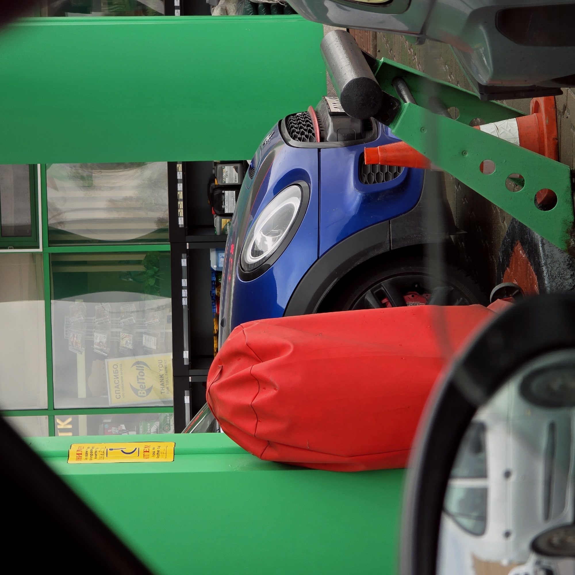 The image captures a scene from inside a vehicle, looking out at a service station or car wash bay. A large, red, crinkled object, possibly a bag or car cover, occupies a significant portion of the foreground. Beyond this, a prominent green vertical structure frames the left side of the view.
A blue car, identifiable as a Mini Cooper by its distinctive headlight and grille design, is parked in the middle ground. Its front-left wheel is visible, revealing a red brake caliper with "WORKS" branding. Two orange traffic cones are positioned around the car, suggesting a designated area or service bay. To the right, part of a green metal structure with circular cutouts is visible, with another orange cone behind it. Behind the blue car, shelves stocked with various bottles, likely automotive fluids, are arranged.
The scene includes a large window reflecting the interior of a building. Prominently visible in the reflection is a yellow sign with text in both Russian and English: "СПАСИБО, ЧТО ОПЛАТИЛИ ПРОЕЗД" and "THANK YOU FOR TOLL PAYMENT," accompanied by the "BelToll" logo. Other partial Russian texts from the OCR include "ВНИМЕ" (Attention), "нефть" (oil), "вам", "прав", "Вните", and "TK". The overall lighting suggests it is daytime. The setting appears to be a commercial service area for vehicles, situated in Дорскі сельскі Савет, Belarus.