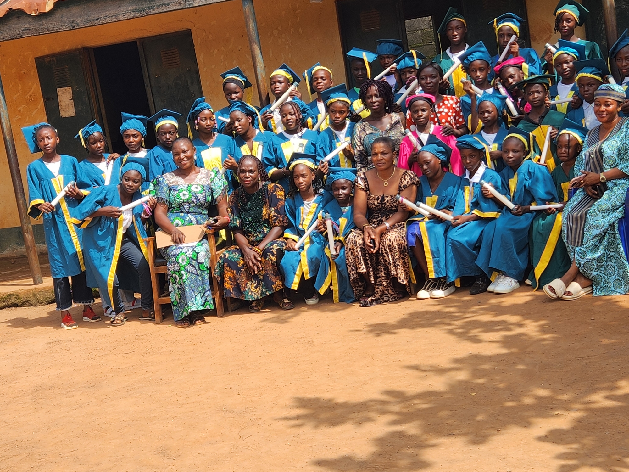 In Sabon Tasha, Nigeria, a group of individuals, mostly young students in graduation attire, are gathered outdoors for a ceremony. The students, a mix of genders, wear matching teal gowns with yellow trim and matching caps, many holding rolled-up certificates. Several adults, presumably teachers or parents, are interspersed among them, also dressed in traditional Nigerian attire. The setting appears to be a schoolyard with a building in the background, featuring yellow walls and dark doorways. The ground is covered in reddish-brown dirt, with shadows of trees cast across it, indicating an outdoor event on a sunny day. The overall atmosphere suggests a graduation or similar celebratory academic occasion.