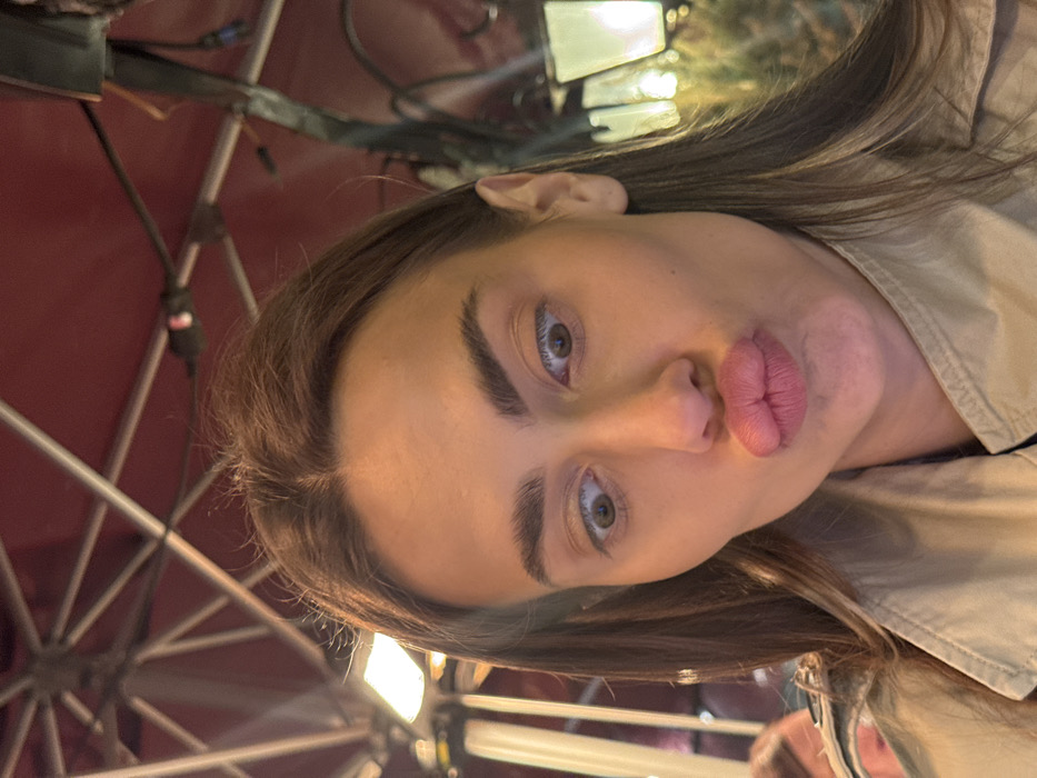 The image displays a close-up, selfie-style portrait of a female individual in the city of Venice, Italy. The subject has long, dark brown hair, prominent dark eyebrows, and light brown eyes that are crossed and directed downwards. Her lips are heavily pursed into a distinct "kissing" or "duck face" expression. She is wearing a light-colored, possibly beige, trench coat. The background indicates an outdoor or semi-outdoor restaurant or cafe setting under low light conditions. Overhead, a red or maroon canopy or awning with visible metal support beams is present. A rectangular, illuminated light fixture is visible on the left, attached to the overhead structure. To the right, a traditional, illuminated street lamp or lantern with a dark frame and glowing panels is partially visible. A blurred, out-of-focus head and shoulder of another person are discernible in the lower-left background.