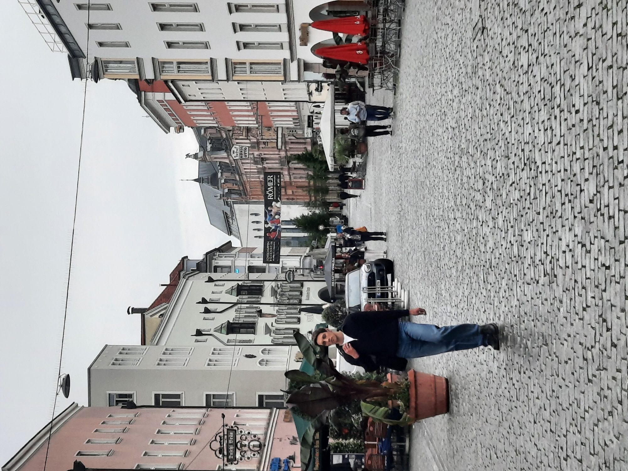 The image depicts a view of a cobblestone street in Rosenheim, Germany. In the foreground, a man is walking towards the camera. He is wearing a dark jacket, blue jeans, and black shoes. He is carrying a dark bag on his left shoulder. To his left, there is a large potted plant with dark foliage. In the background, there are buildings lining the street with various businesses. Signs visible on the buildings include "ES CAFFE VENEZIA" and "RÖMER". There are also tables and chairs set up outside some of the establishments, with red umbrellas visible. Several other individuals are seen walking in the distance, and a dark-colored car is parked on the street. The sky is overcast, suggesting a cloudy day.