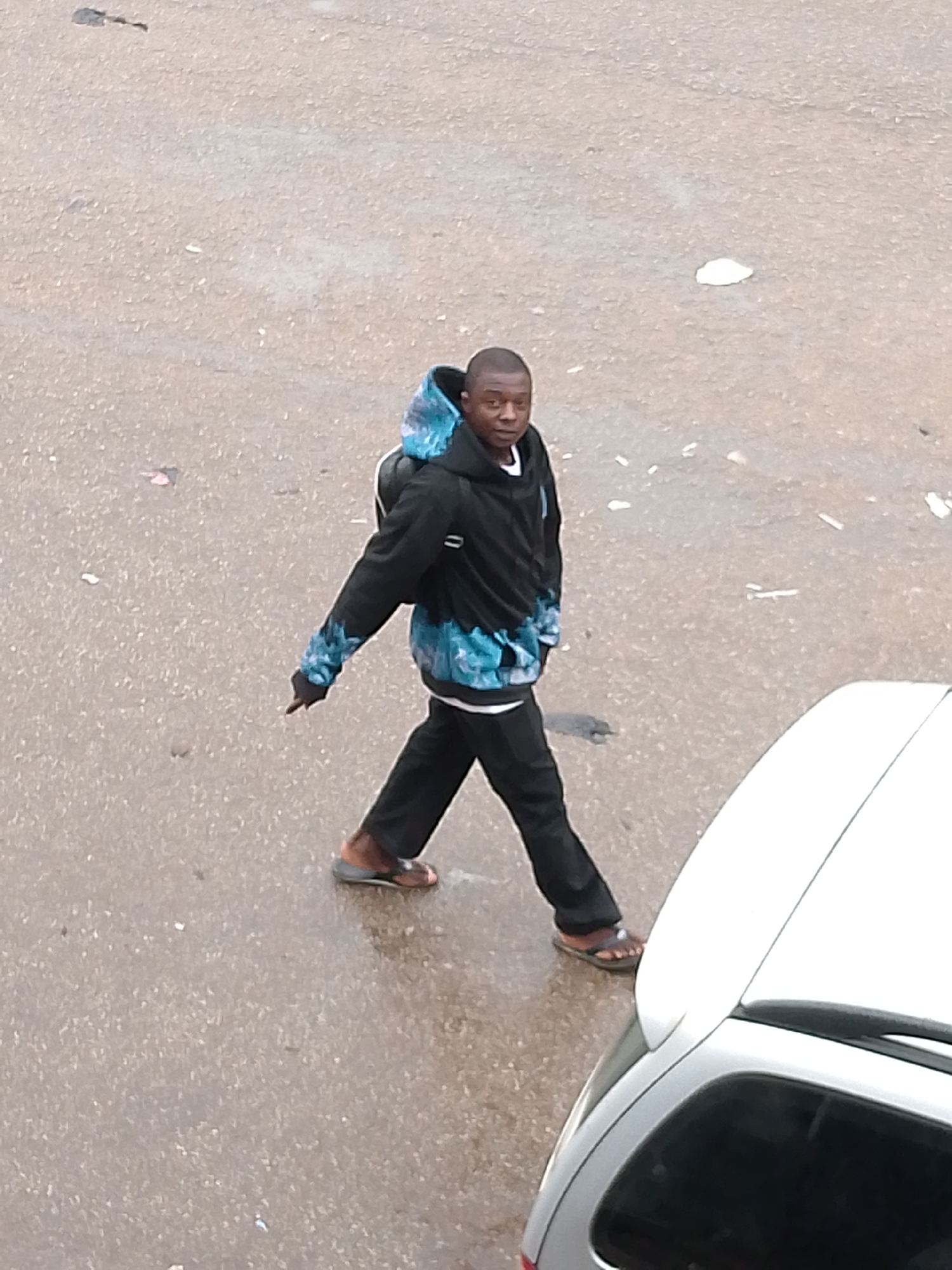 An overhead shot captures a young man walking on a wet, paved surface, likely a street or parking lot, in Bukuru, Nigeria. He is seen in mid-stride, moving from the upper left towards the lower right of the frame, with his gaze directed towards the viewer.
The man, who appears to be of African descent, wears a black hooded sweatshirt with a distinctive blue and white tie-dye pattern on the sleeves and the lower part of the torso. He has a black backpack on and is dressed in black trousers and brown flip-flops or sandals. His left arm is extended, with his index finger pointing downwards or forwards.
The ground beneath him is a textured grey, appearing damp or wet, possibly from recent rain or a damp environment. Small pieces of debris, including white and darker specks, are scattered across the pavement. In the lower right corner, the rear section of a white car with a dark window is visible. The lighting is diffused, suggesting an overcast day. No text is visible in the image.