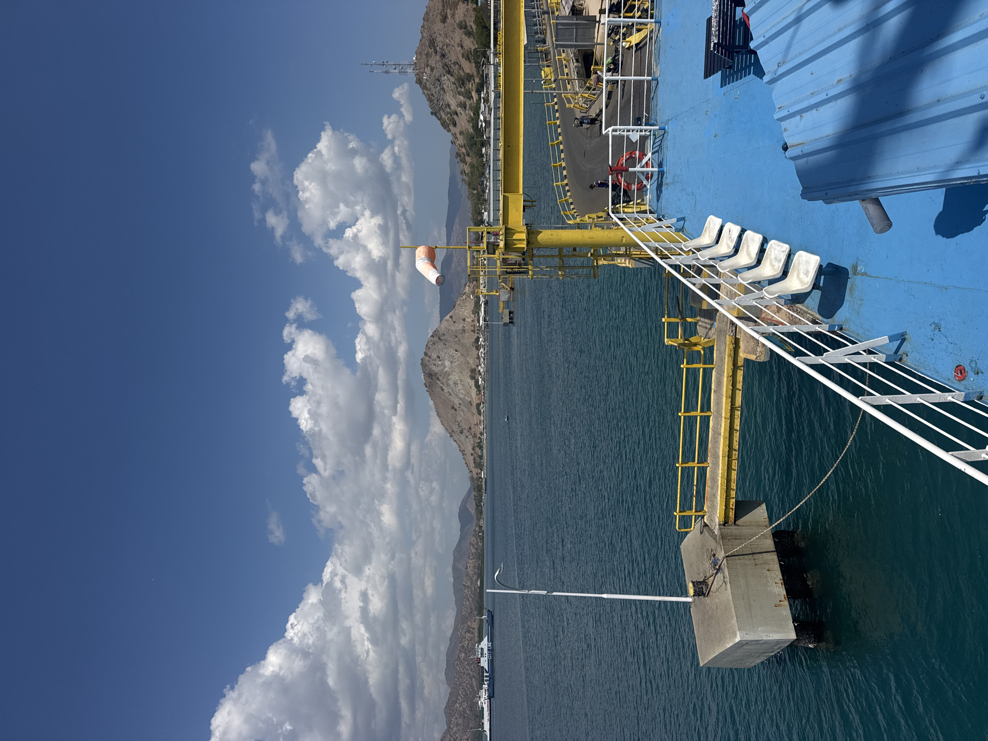 The image depicts a sunny day at a port in Upt Tambak Sari, Indonesia. In the foreground, the blue deck of a ferry is visible, lined with white railings. Several white plastic seats are arranged along the railing, facing the water. To the left, a concrete pier extends into the turquoise water, connected to a yellow walkway with railings. A white lamp post stands on the pier. In the midground, the calm blue sea stretches towards a coastline dotted with brown, arid hills. Several ferries are anchored or moored in the distance. A tall yellow structure with a windsock is prominent on the right side of the image, near a roadway with a few people and motorcycles. The sky is a clear blue, with scattered white clouds. A communication tower is visible on one of the hills. The overall scene suggests a functioning port with infrastructure for ferry services in a coastal, somewhat dry environment.