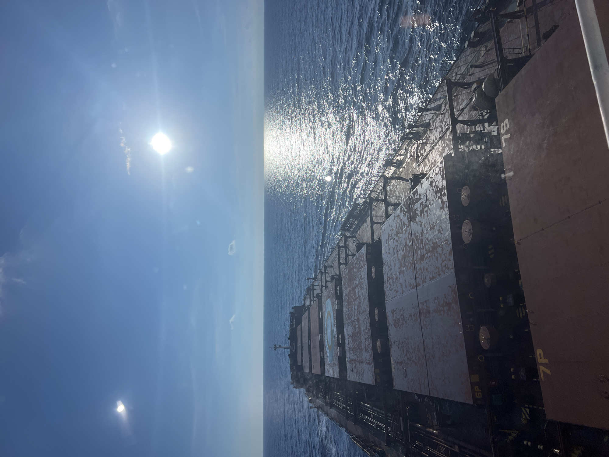 The image shows a cargo ship sailing on a calm, blue ocean under a bright, sunny sky. The sun is high in the sky, casting a strong glare on the water and creating a shimmering reflection. The deck of the ship is covered with large, flat cargo hatches, some of which have numbers stenciled on them, such as "6P" and "7P". The ship's superstructure, including a mast and railings, is visible towards the stern. The horizon is clearly defined, with the ocean meeting the sky in a seamless line. The overall impression is one of a peaceful voyage at sea.
